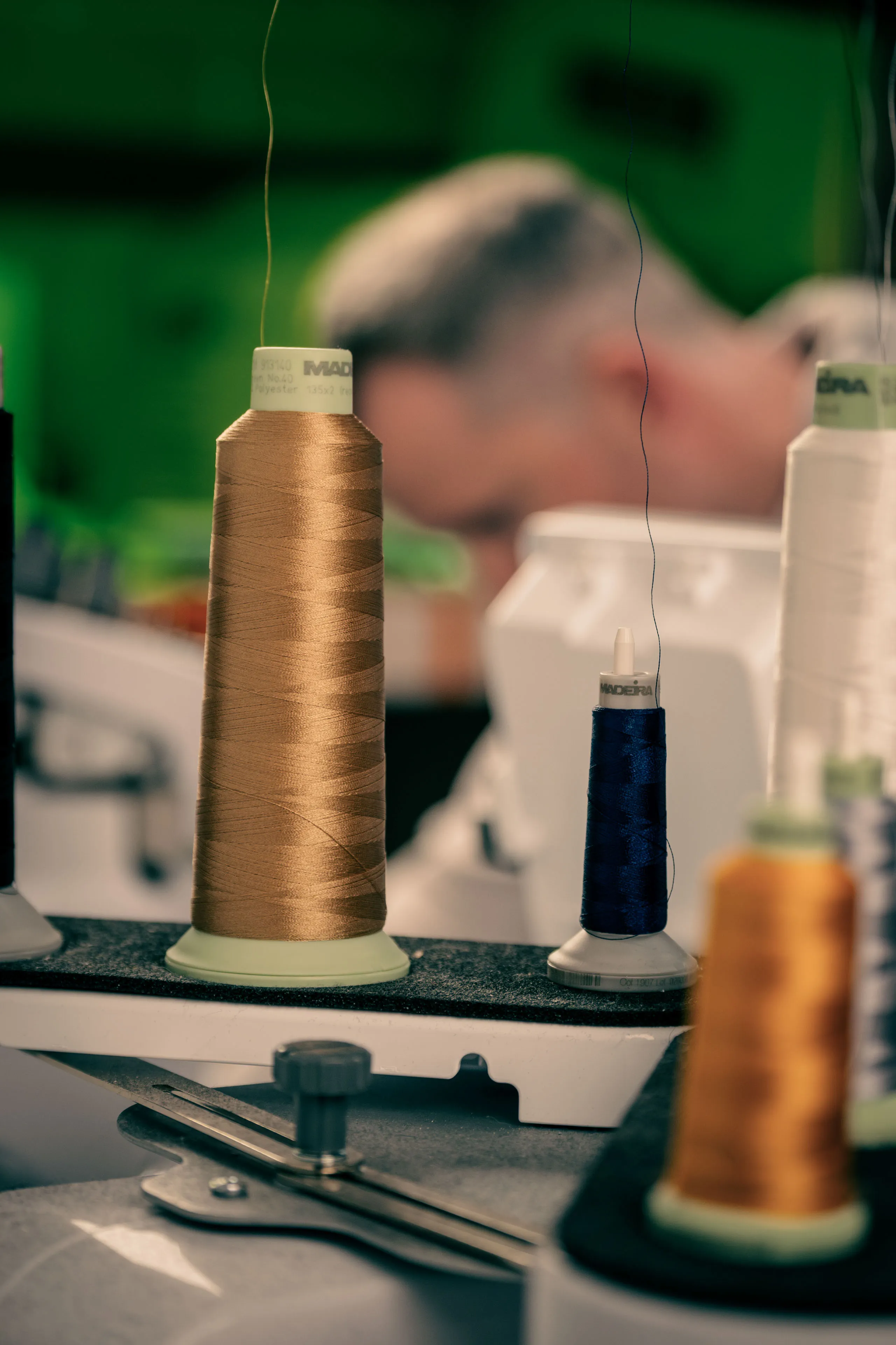 Close-up of spools of brown, blue, and orange thread in a sewing machine, with a blurred figure in the background, conveying focus and craftsmanship.