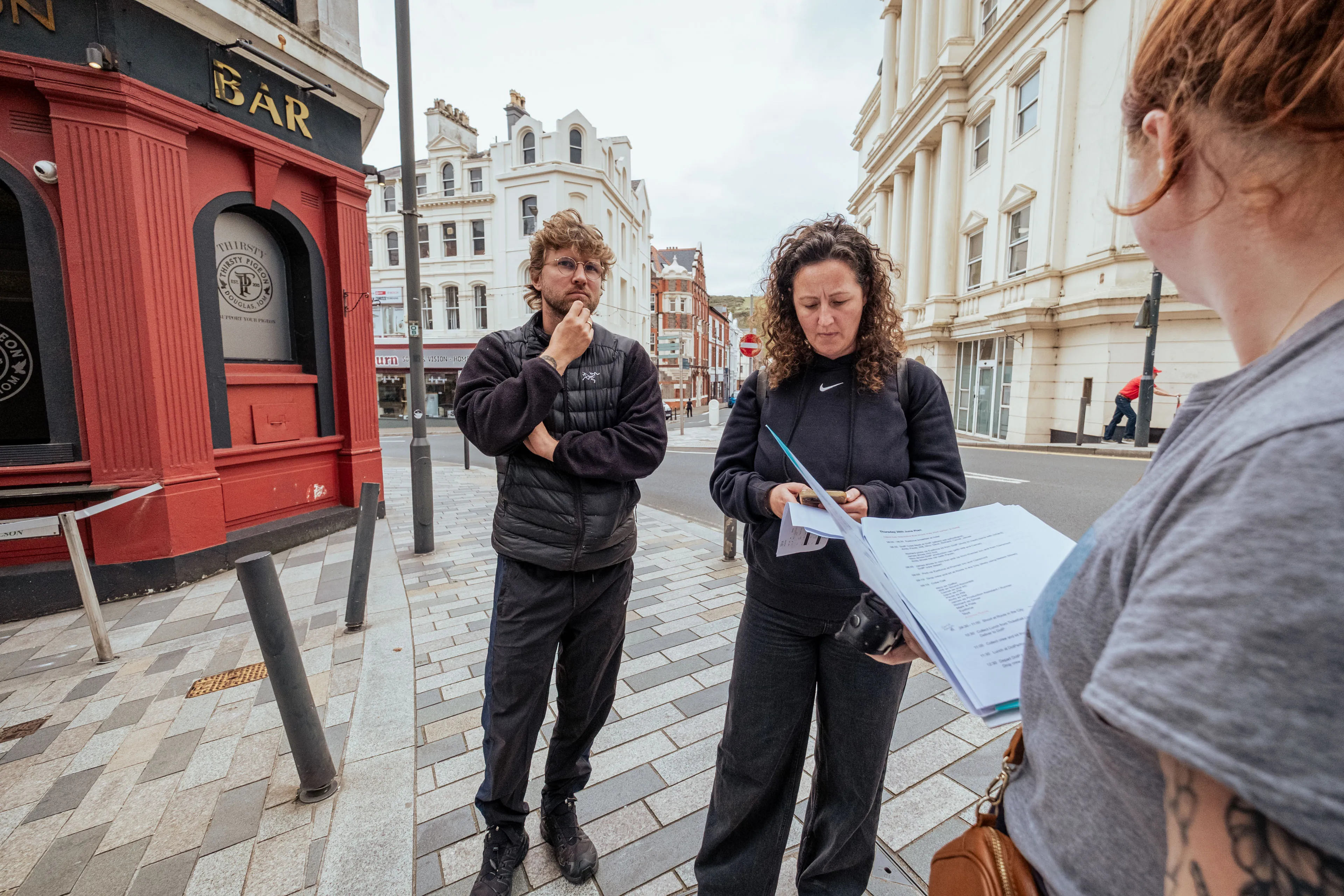 Three people at a street corner, two reading papers with focused expressions. A red building labeled "BAR" in the background. Urban setting, overcast sky.