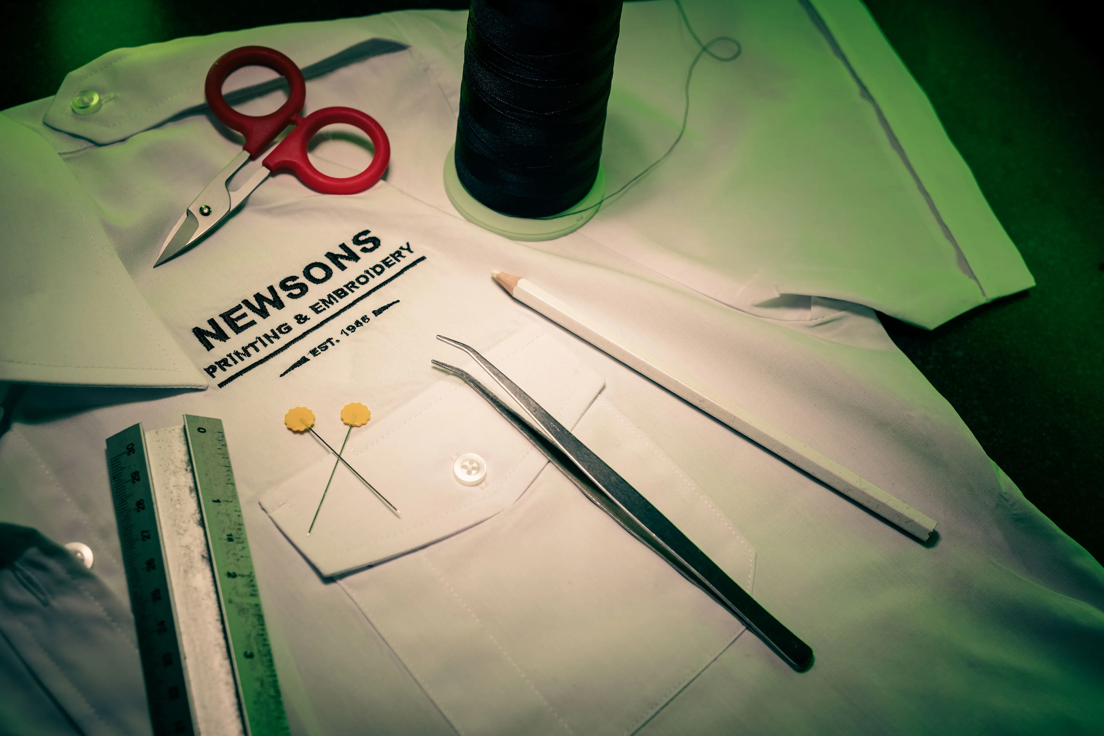 A neatly arranged white shirt labeled "Newsons Printing & Embroidery" with red scissors, black thread, tweezers, sewing pencils, pins, and a ruler nearby.
