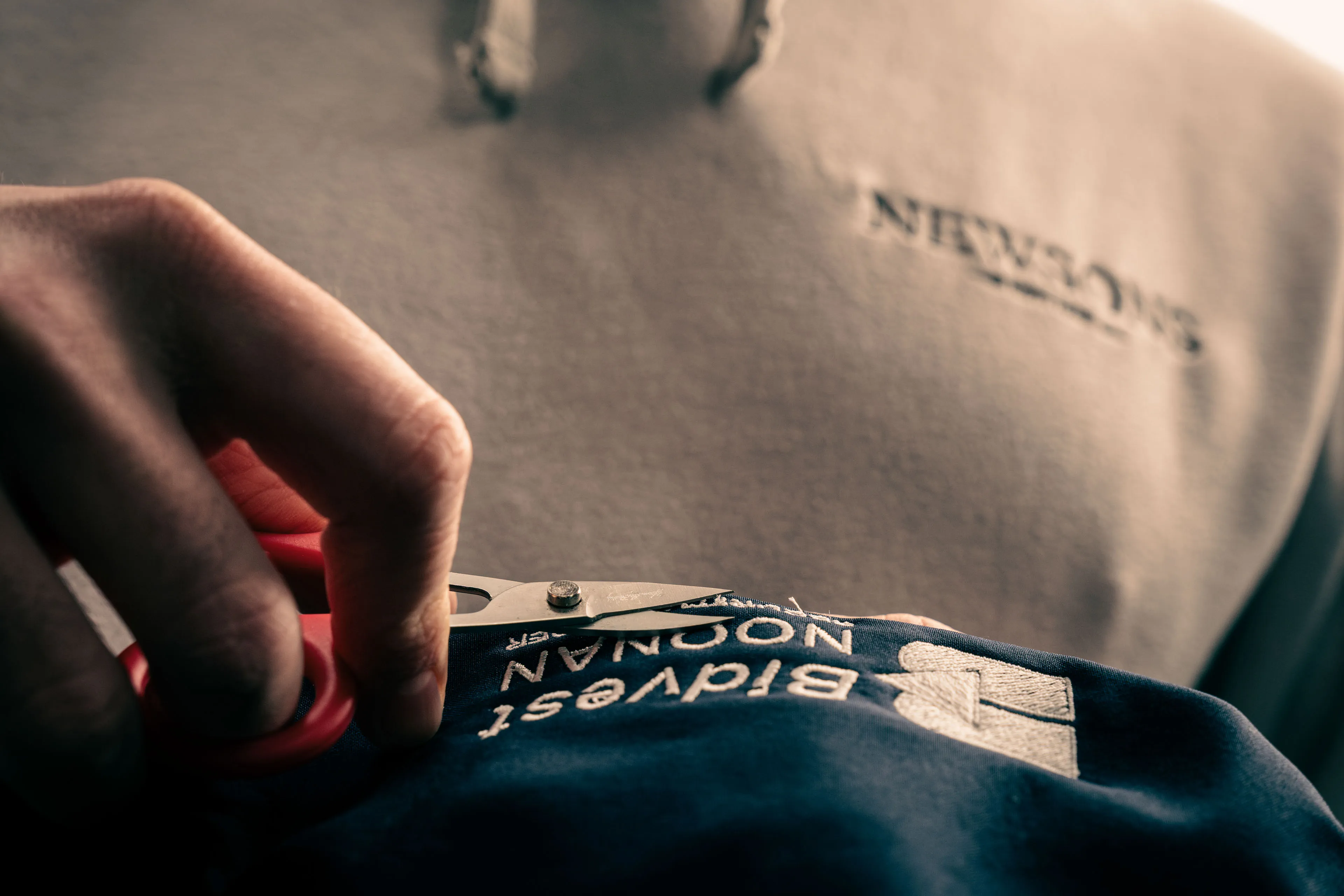 Close-up of a person using red scissors to cut a blue fabric label. The background is blurred, creating a focused and intimate atmosphere.