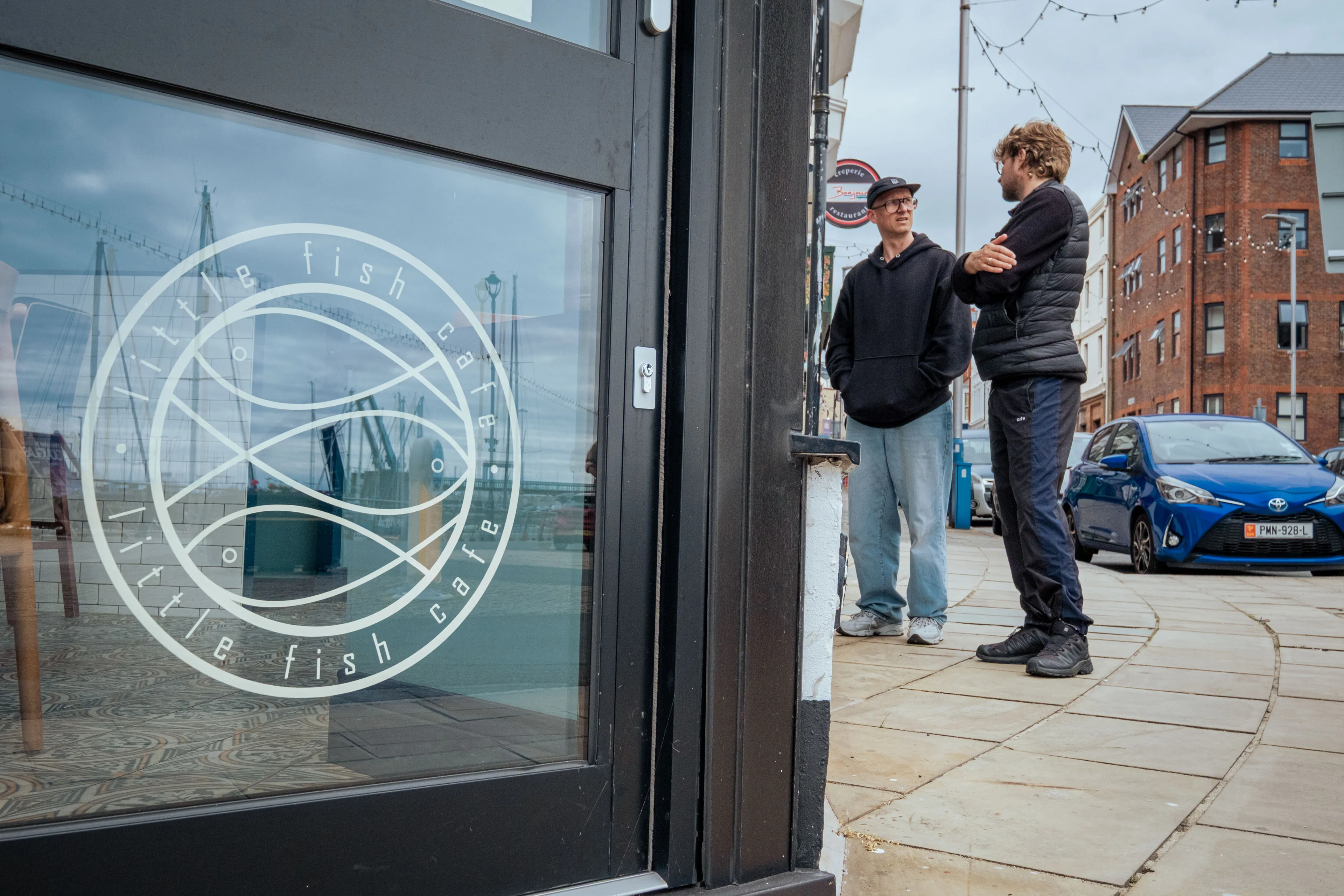 Street scene outside Little Fish Cafe. Two people converse casually on a sidewalk beside a modern black door with the cafe's circular logo. Overcast sky.