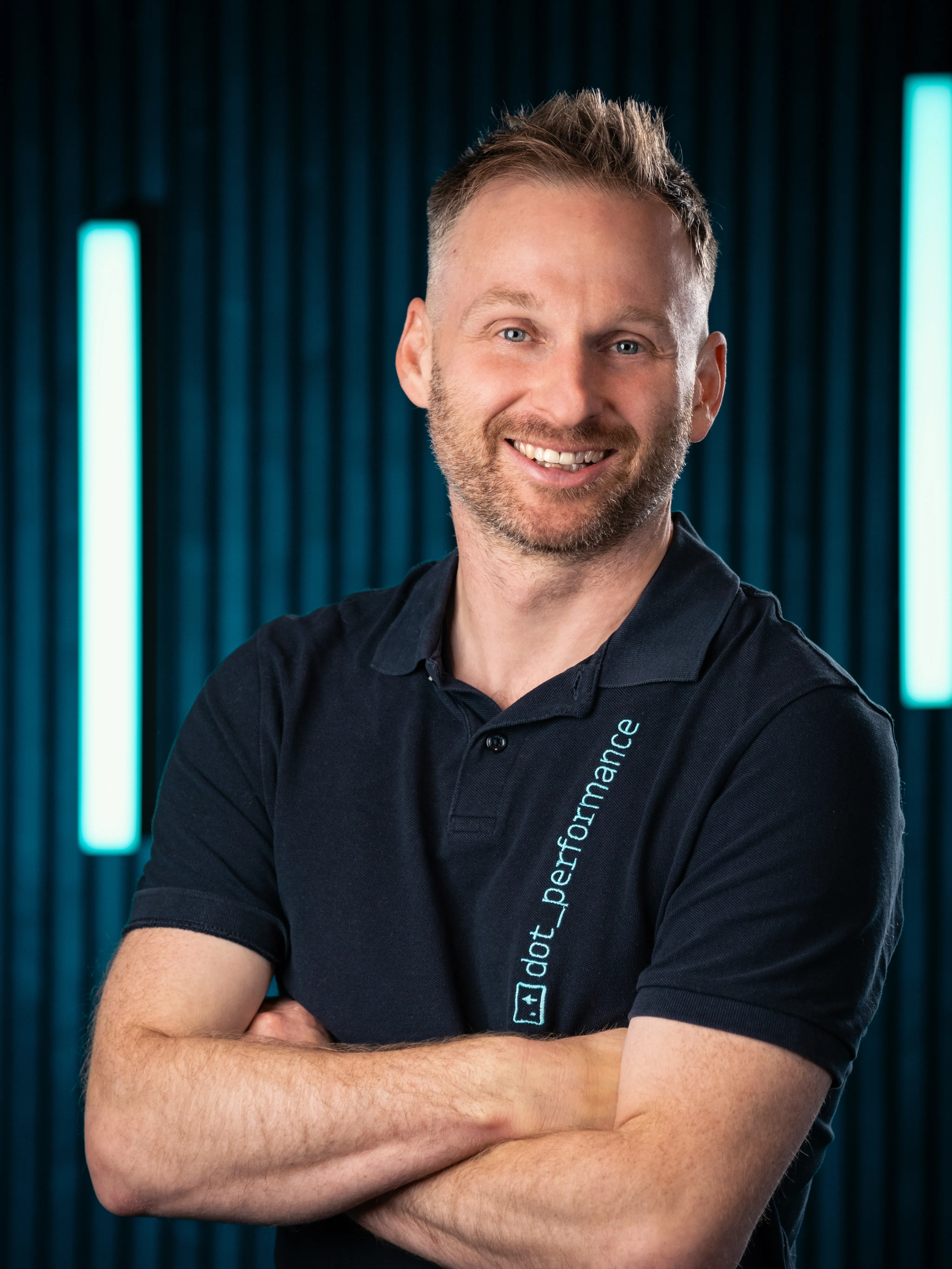 Headshot of John Beaty, head of systems at Dot Performance. He is seen with his arms crossed, smiling, wearing a navy polo shirt with dot performance branding, set against a dark background will neon blue lights.