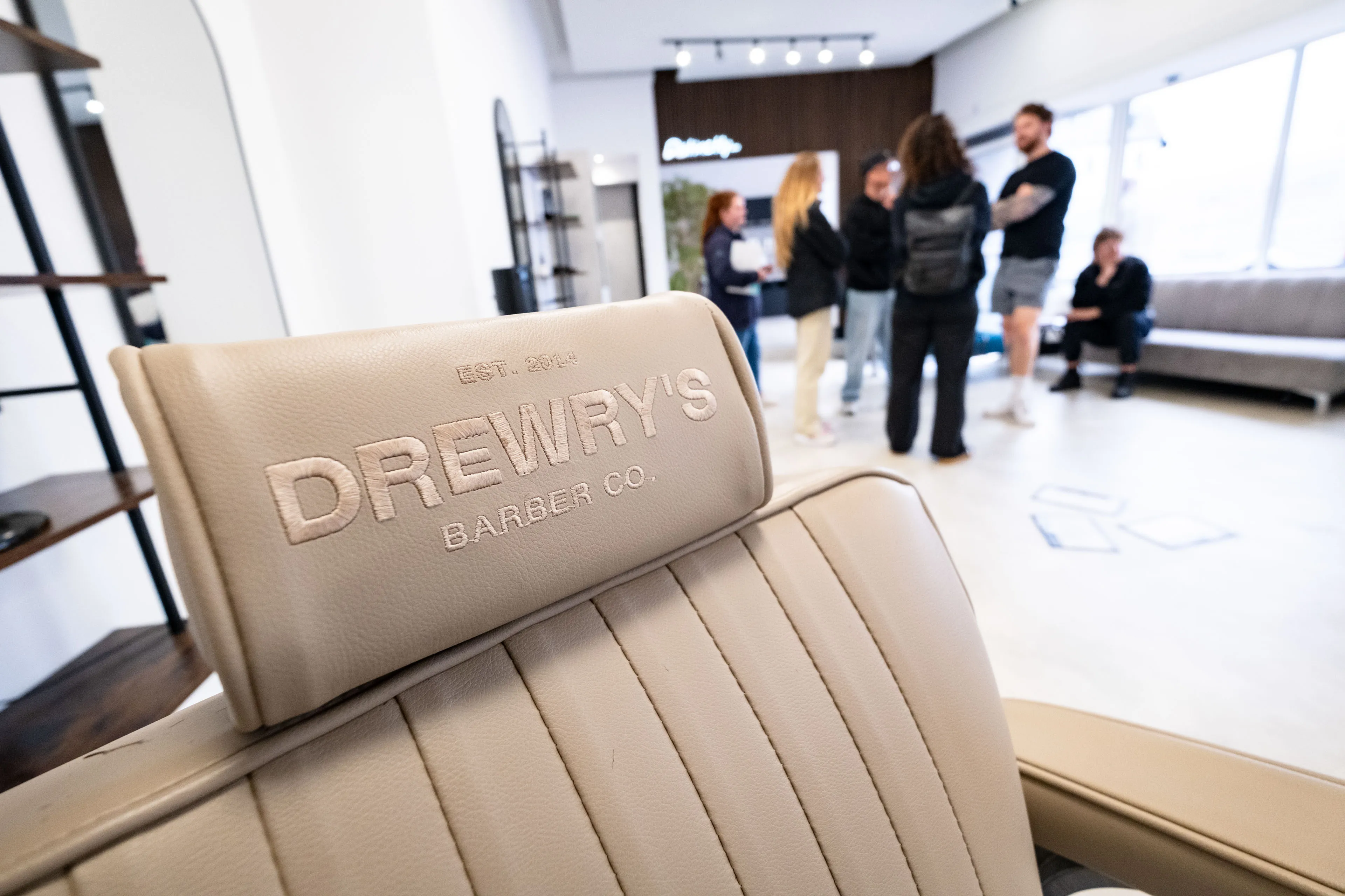 Close-up of a tan barber chair with "DREWRY'S BARBER CO." embossed. In the background, a group of people stand talking in a modern, bright barber shop.