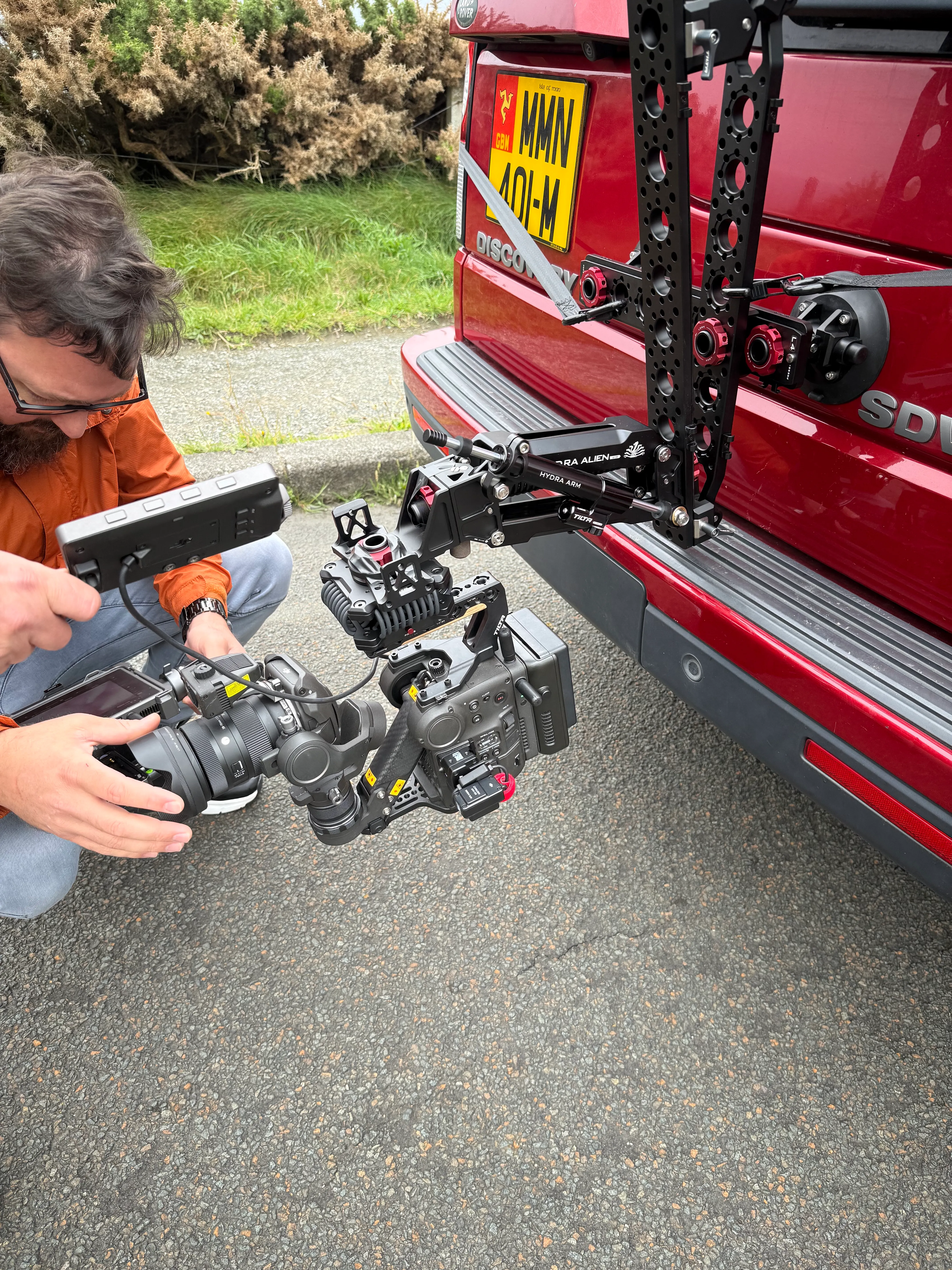 A person with a beard adjusts a professional camera on a red SUV's rear rig, capturing a dynamic setup. The scene conveys a technical and creative focus.
