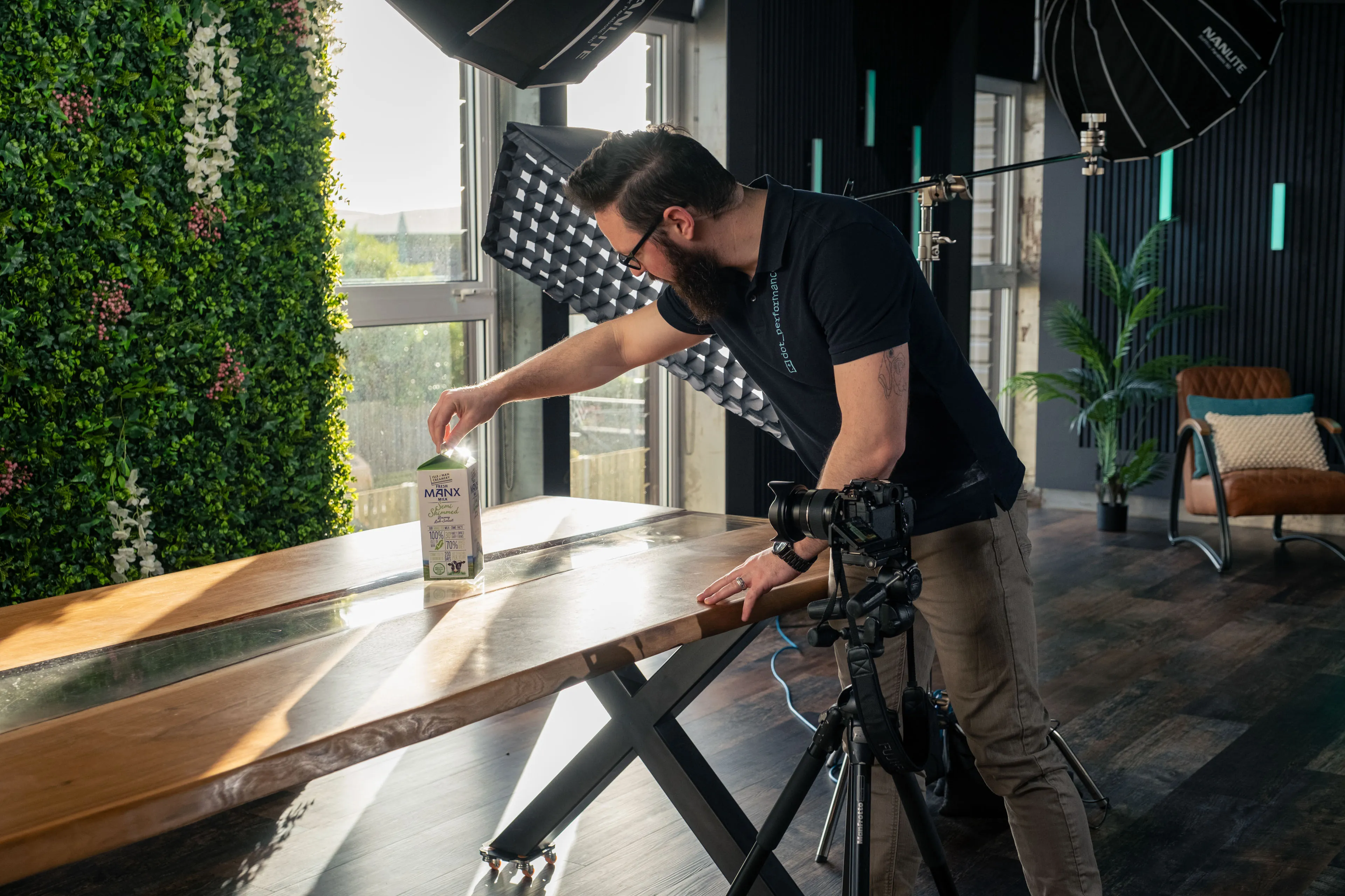 A bearded photographer adjusts a MANX product carton on a glossy wooden table during a studio product shoot with softbox lighting.