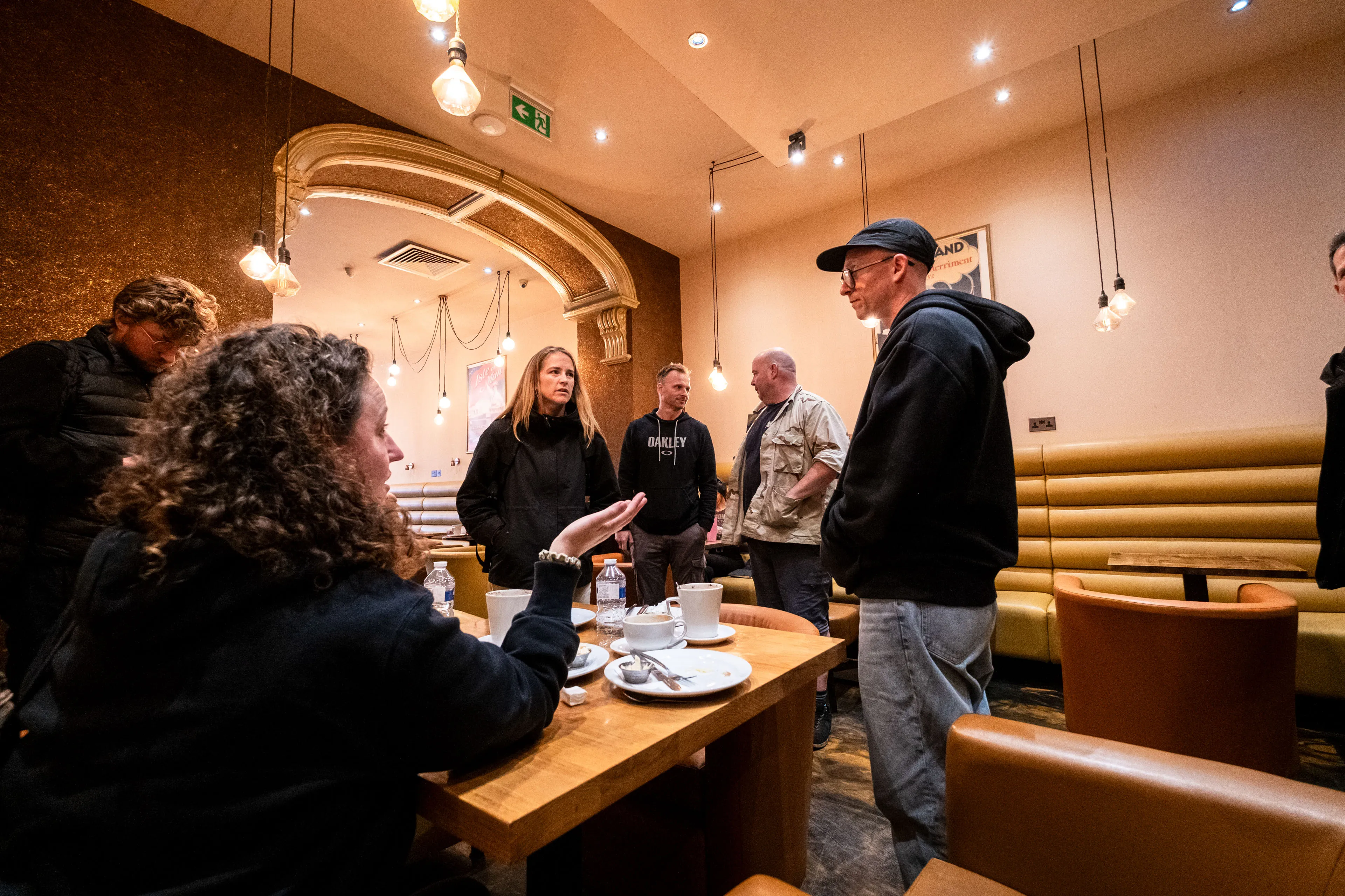 A group of people are gathered in a cozy cafe with warm lighting and arch decor. They seem engaged in a conversation, creating a friendly atmosphere.