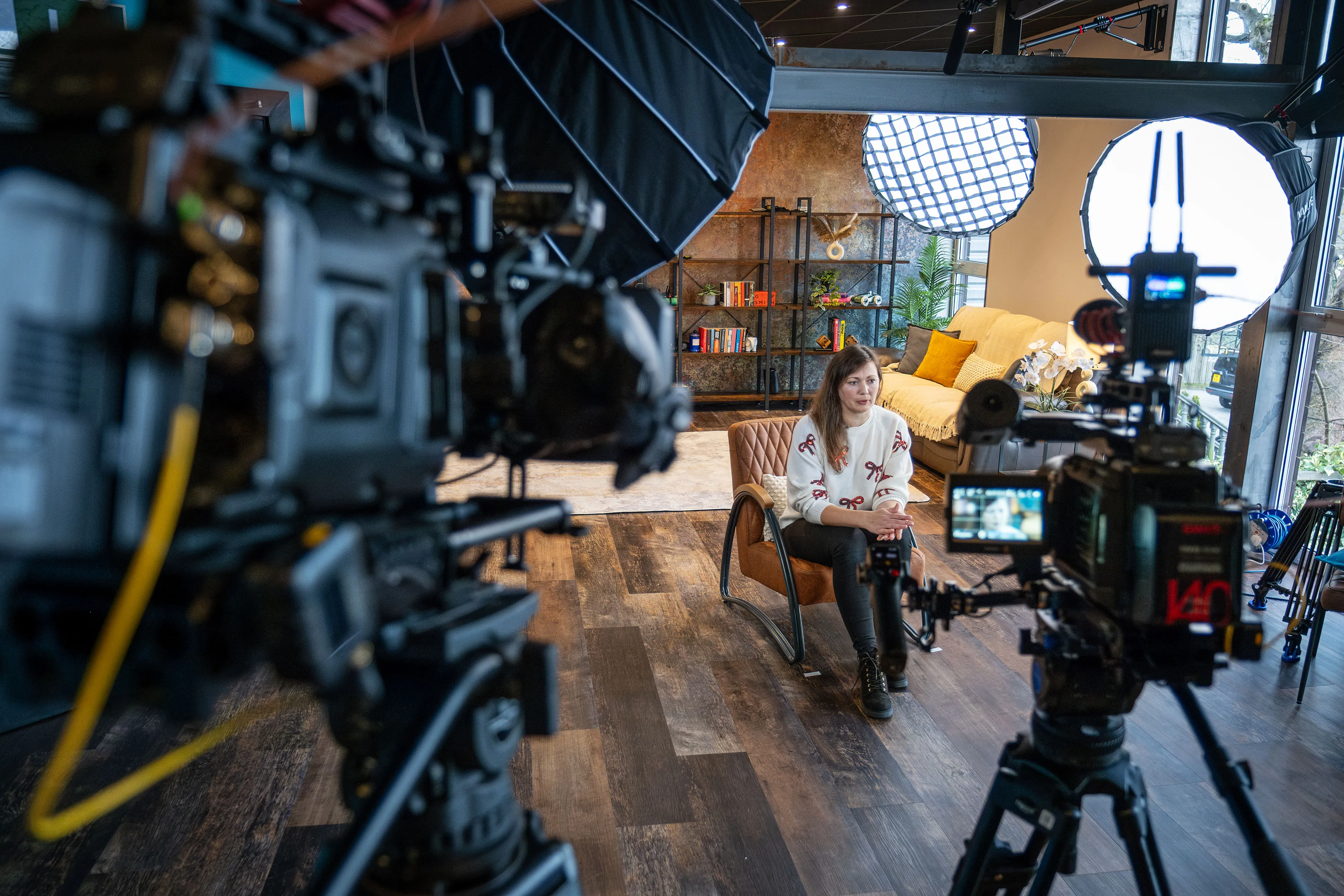 A woman sits on a chair in a well-lit studio, surrounded by professional cameras and lights. She appears focused and calm. A bookshelf and sofa are in the background.