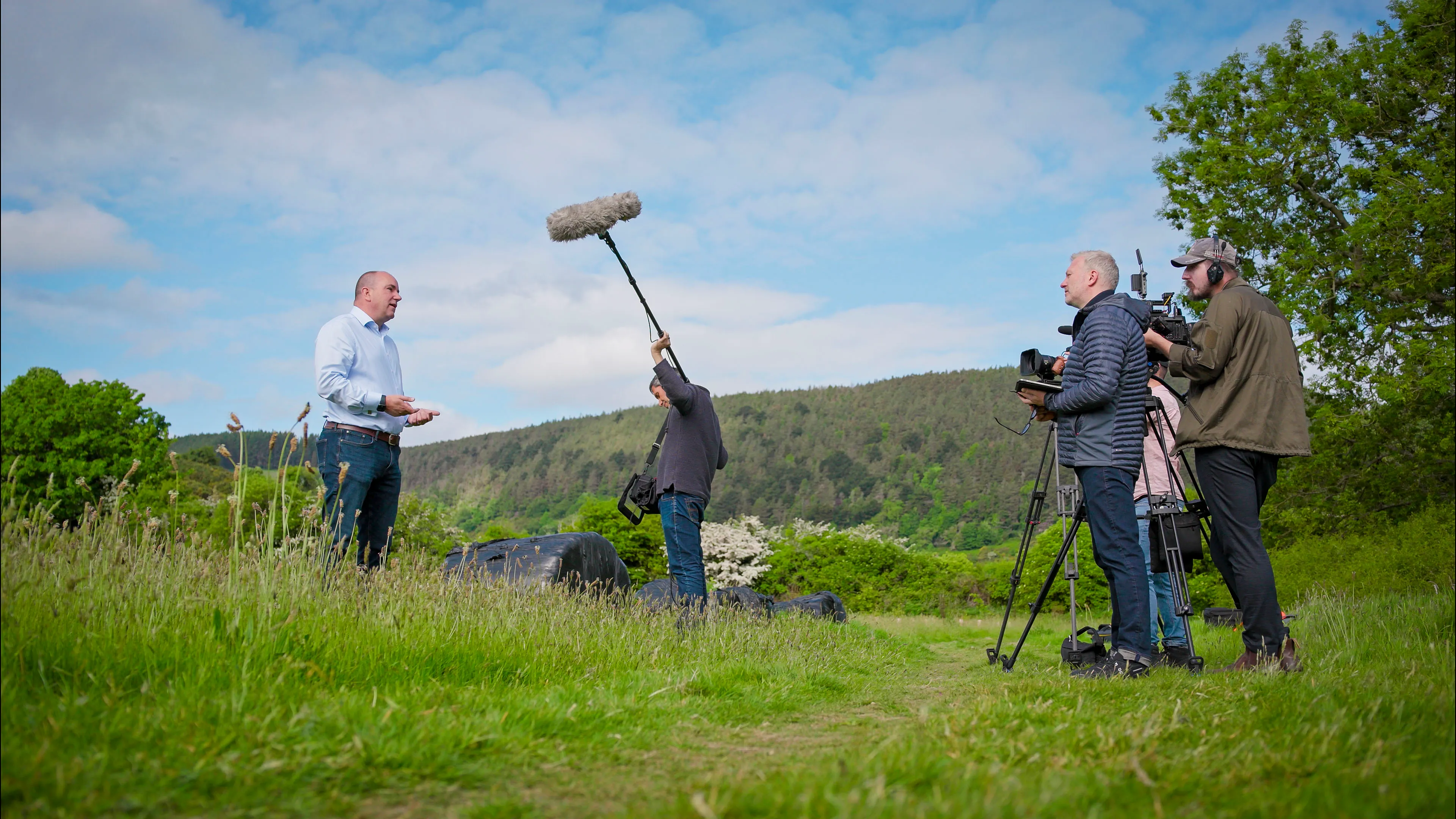 A man is being filmed outdoors on a grassy path with a boom microphone overhead. Three crew members with cameras focus on capturing the scene.