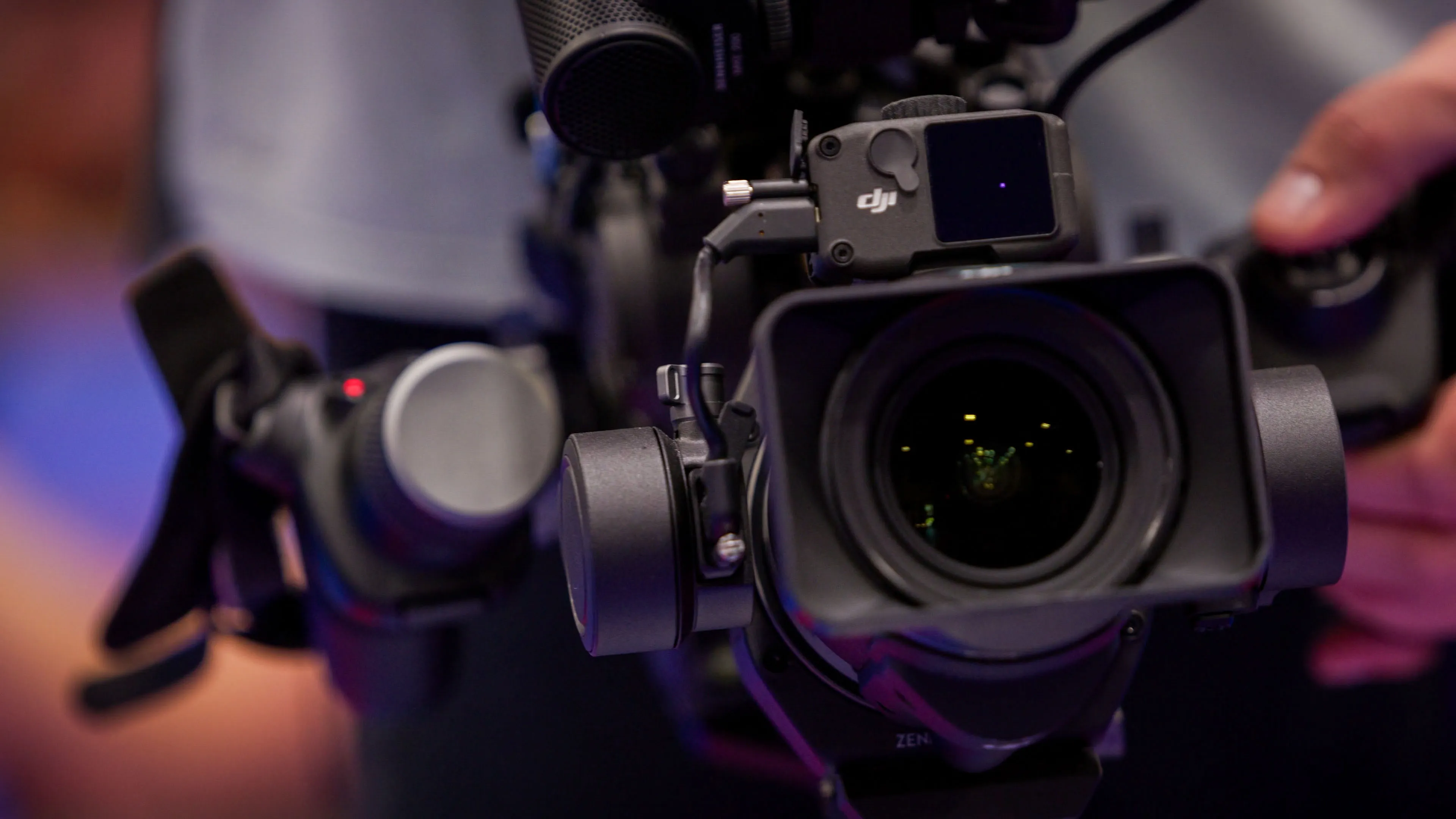 Close-up of a professional camera being held, focused on the lens and gimbal, against a blurred background. The mood is technical and focused.