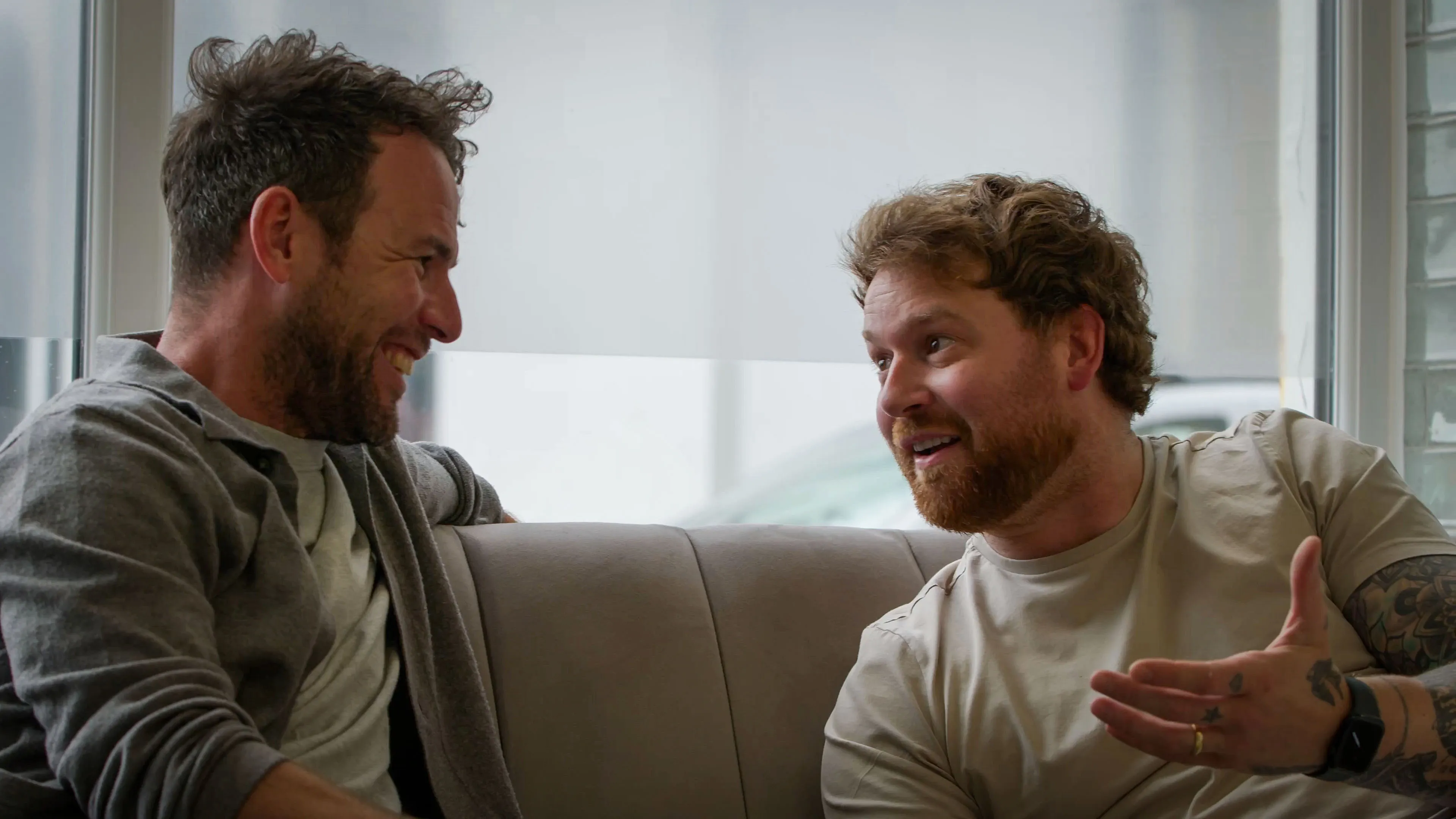 Two men sit on a couch, engaging in lively conversation. Both are smiling, expressing warmth and camaraderie in a casual setting with soft lighting.