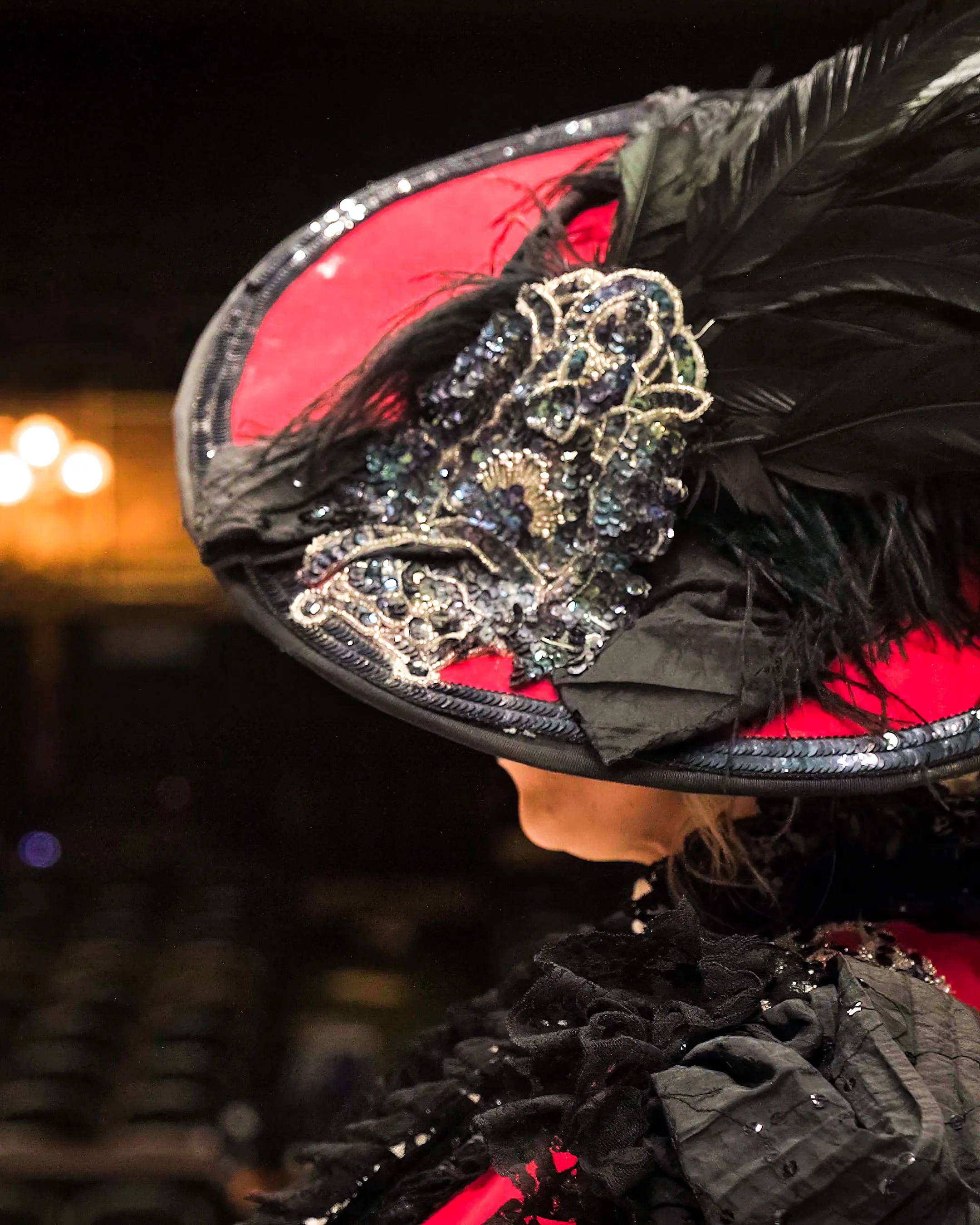 A woman wearing an ornate red and black hat with feathers and intricate beadwork. The background is a dimly lit theater, suggesting a dramatic or theatrical setting.