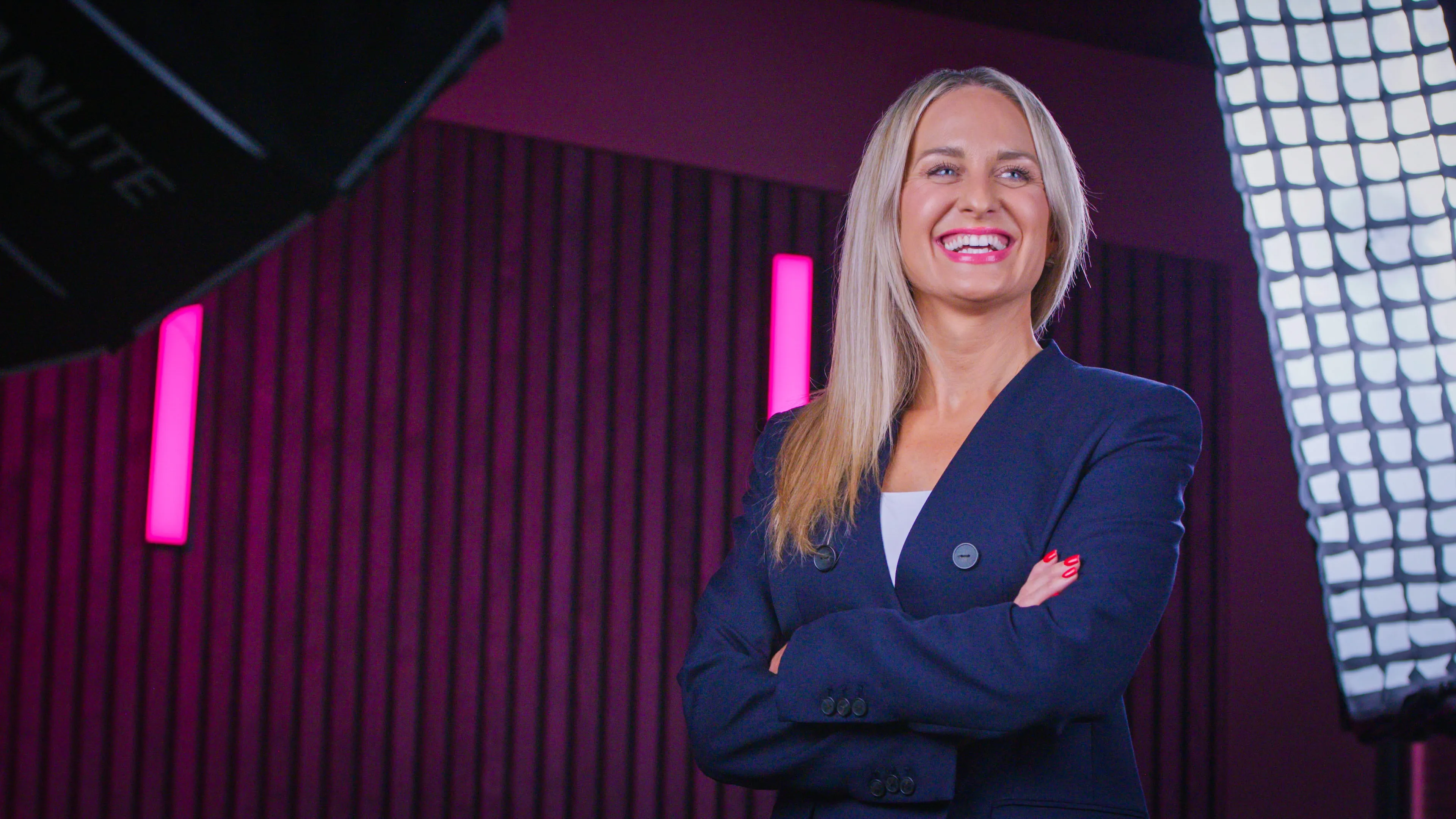 A smiling woman in a navy blazer stands confidently with arms crossed against a pink-striped background, flanked by bright studio lights.