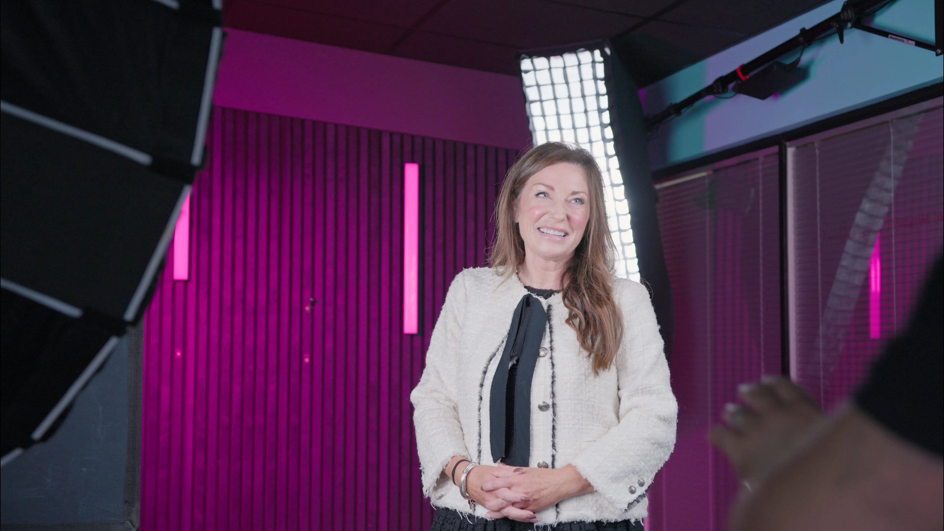 Smiling woman in an off-white jacket stands in a studio with soft pink lighting and professional equipment, conveying a warm, professional ambiance.