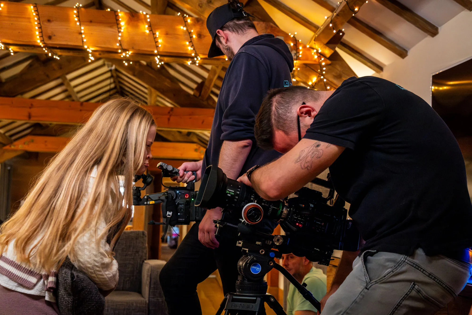 A cozy filming scene in a warmly lit wooden cabin. Two men with cameras and a girl with long blonde hair  pulling faces into the camera. Creating a collaborative, creative atmosphere.