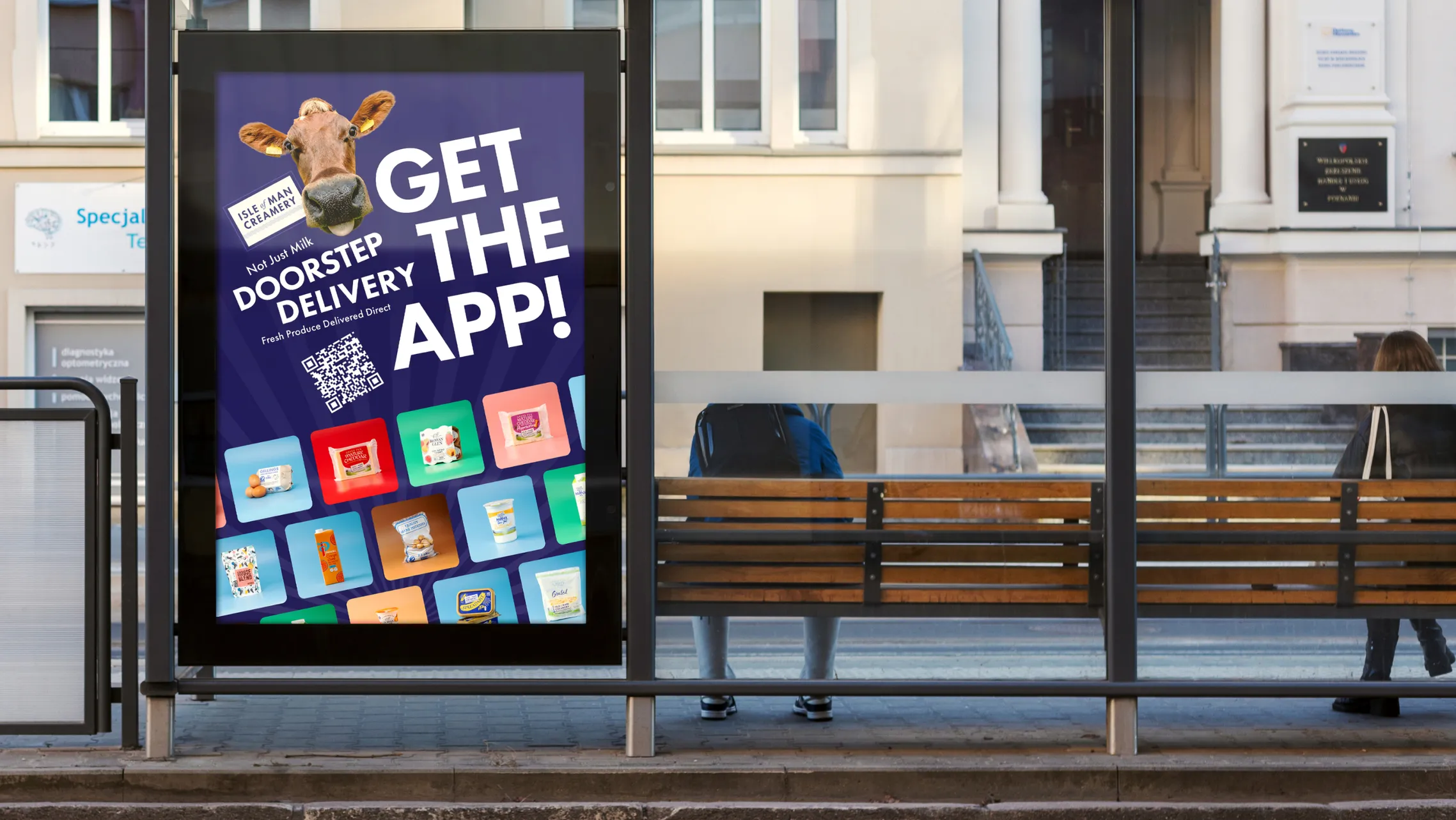 A daytime bus shelter displaying an Isle of Man Creamery "Get the App – Doorstep Delivery" poster, featuring a close-up of a cow's face, a QR code, and a grid of product images, with pedestrians and a building visible in the background