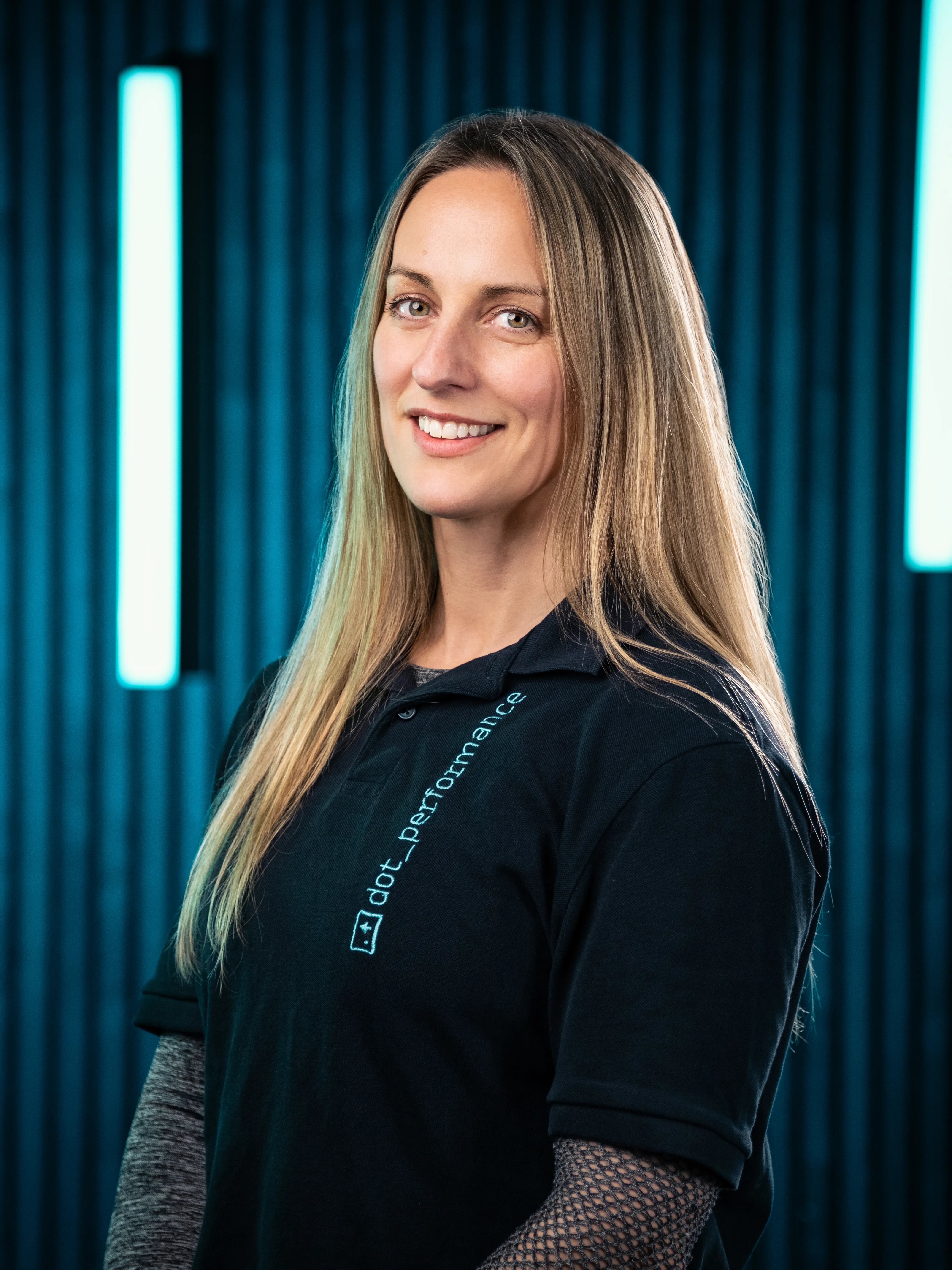 Rachel, customer services manager at Dot Performance, smiling as she poses for headshot wearing branded polo shirt, against dark background with blue neon lights.