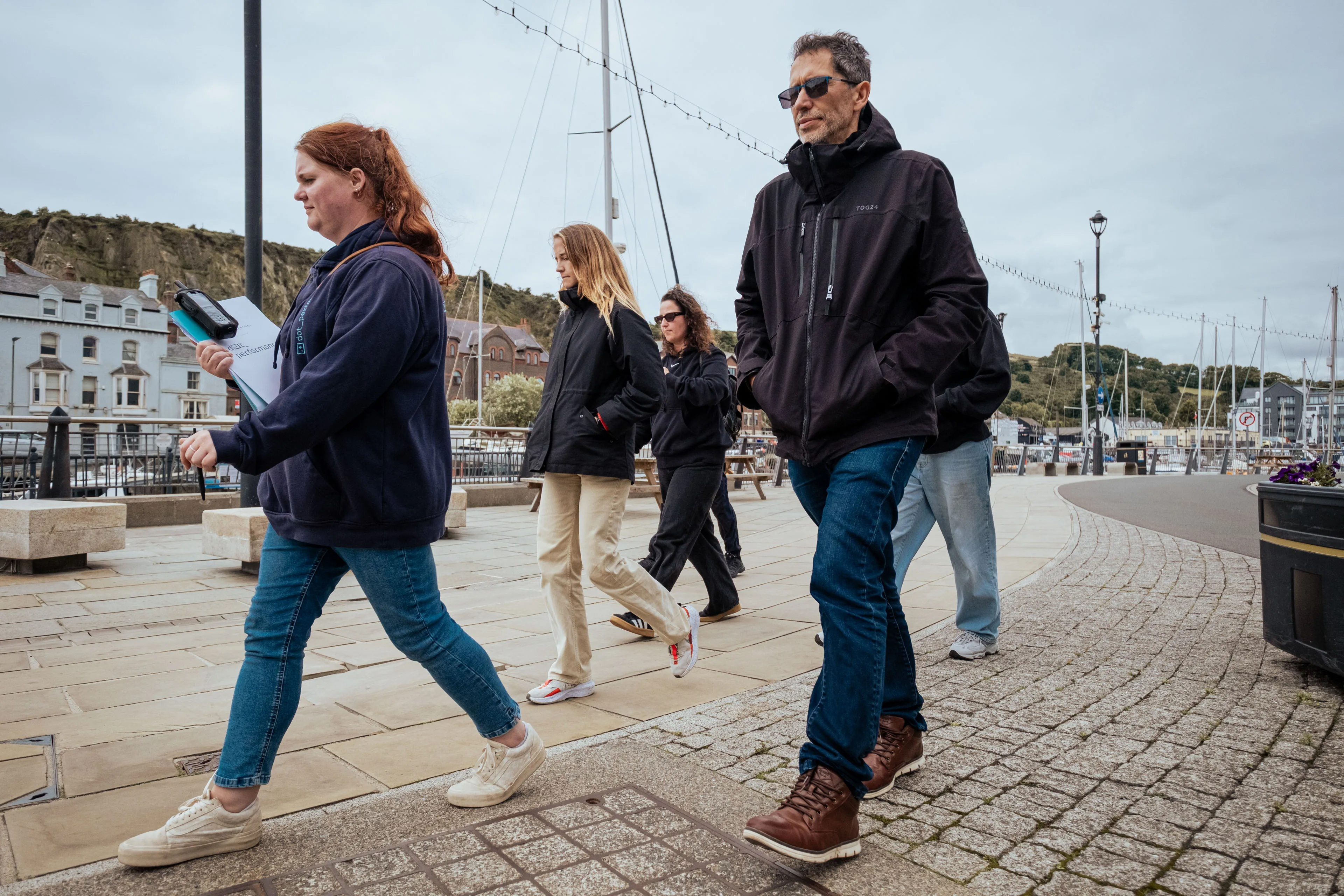 A group of people walks along a marina, dressed in casual jackets and jeans. The overcast sky and boats create a relaxed, seaside atmosphere.