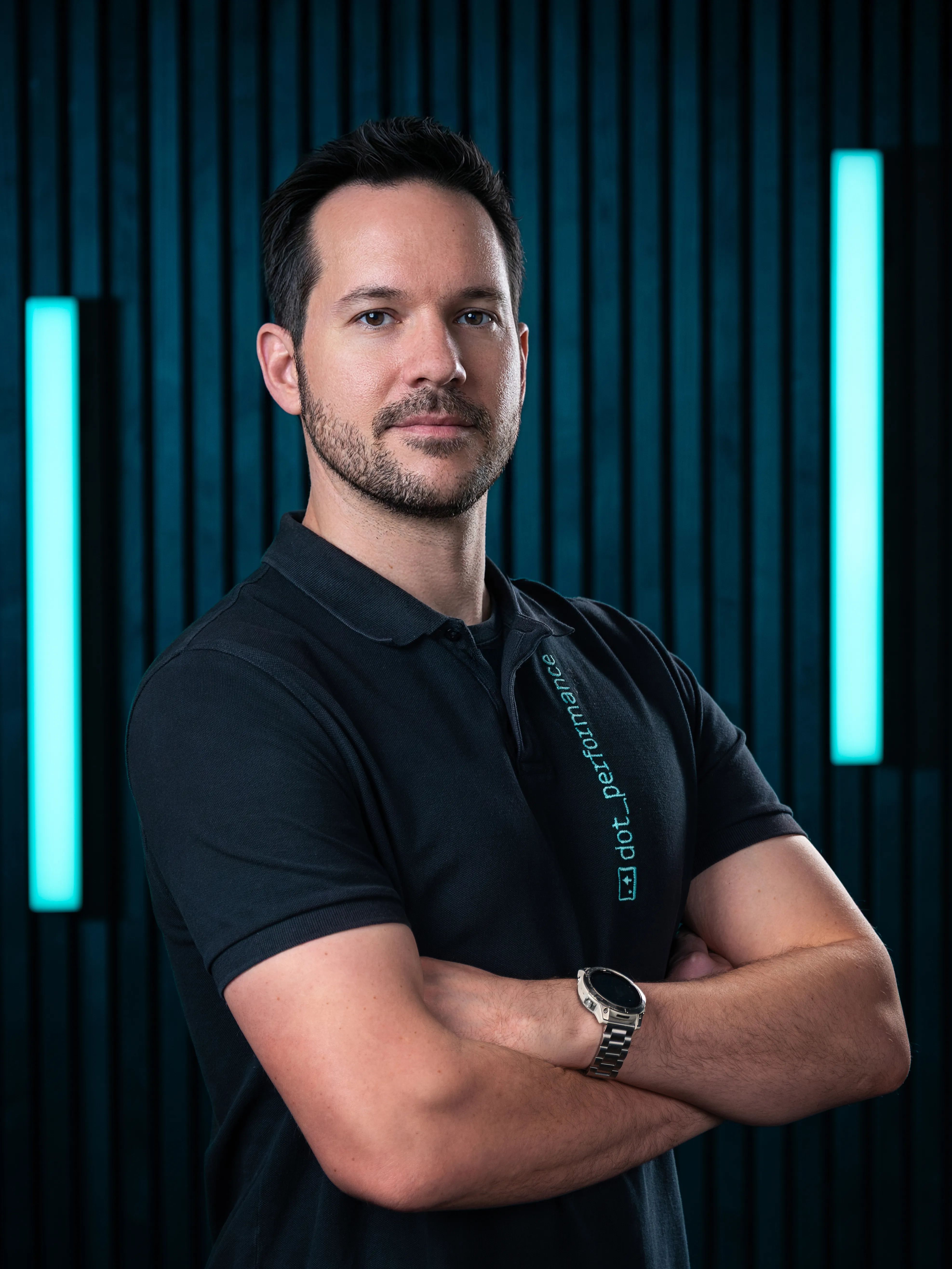 Dave, senior developer at Dot Performance, smiling with crossed arms as he poses for headshot wearing branded polo shirt, against dark background with blue neon lights.