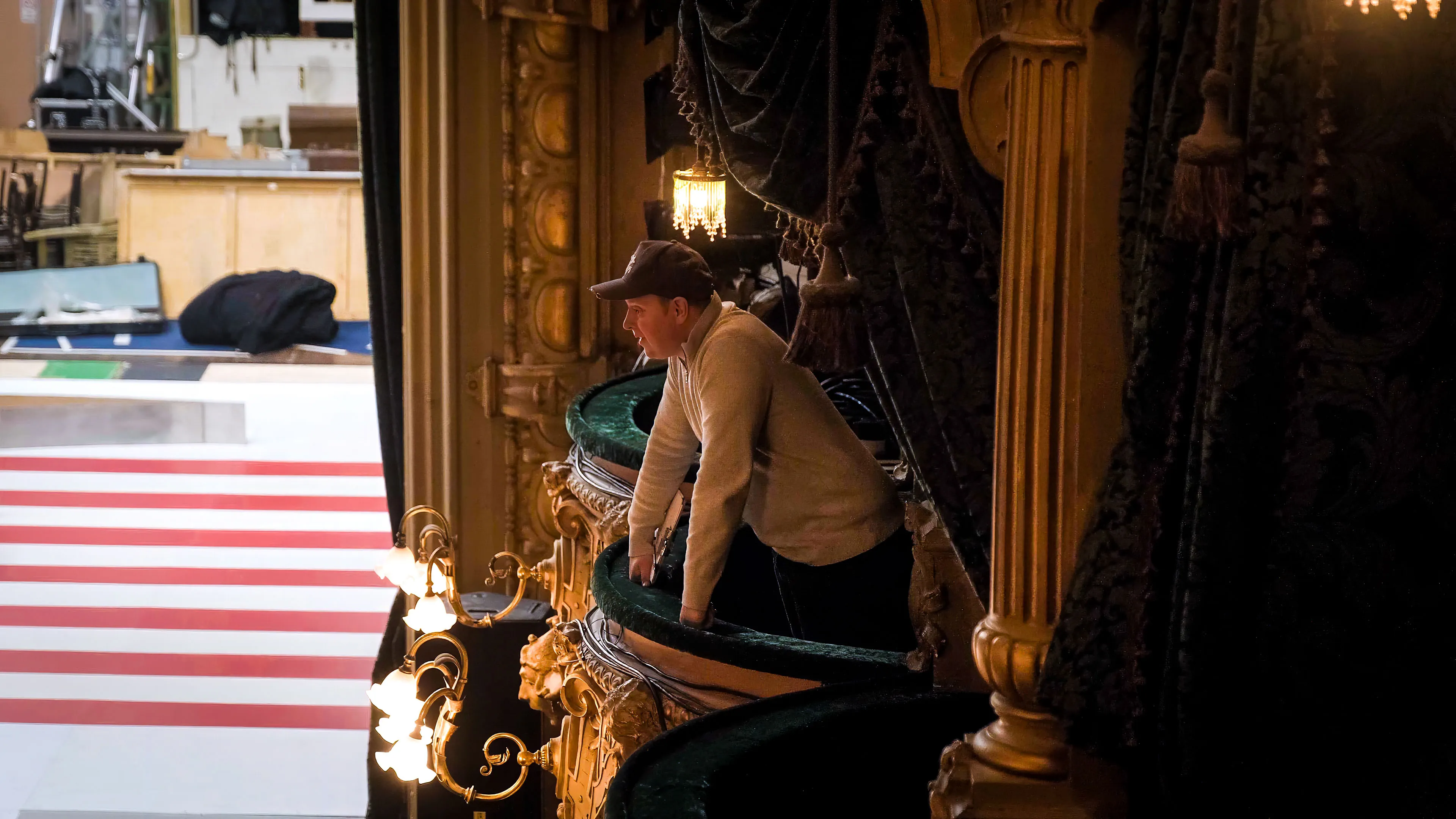 A person in a cap and sweater leans on an ornate theater balcony, observing the stage below. The setting is elegant, with dim lighting and vintage decor.