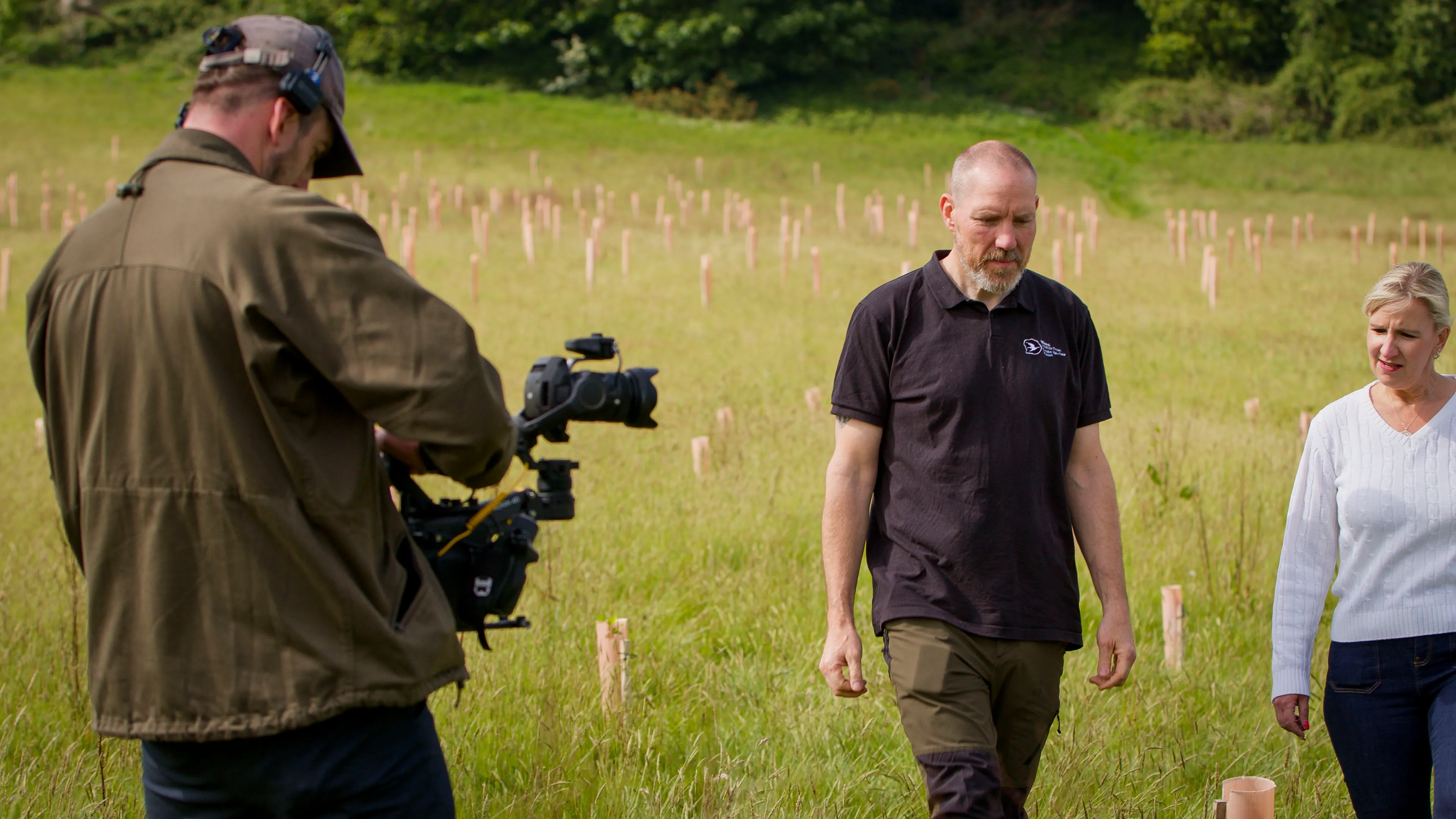 A cameraman films two people walking in a grassy field dotted with tree saplings. The setting is serene with lush greenery and a focus on nature.