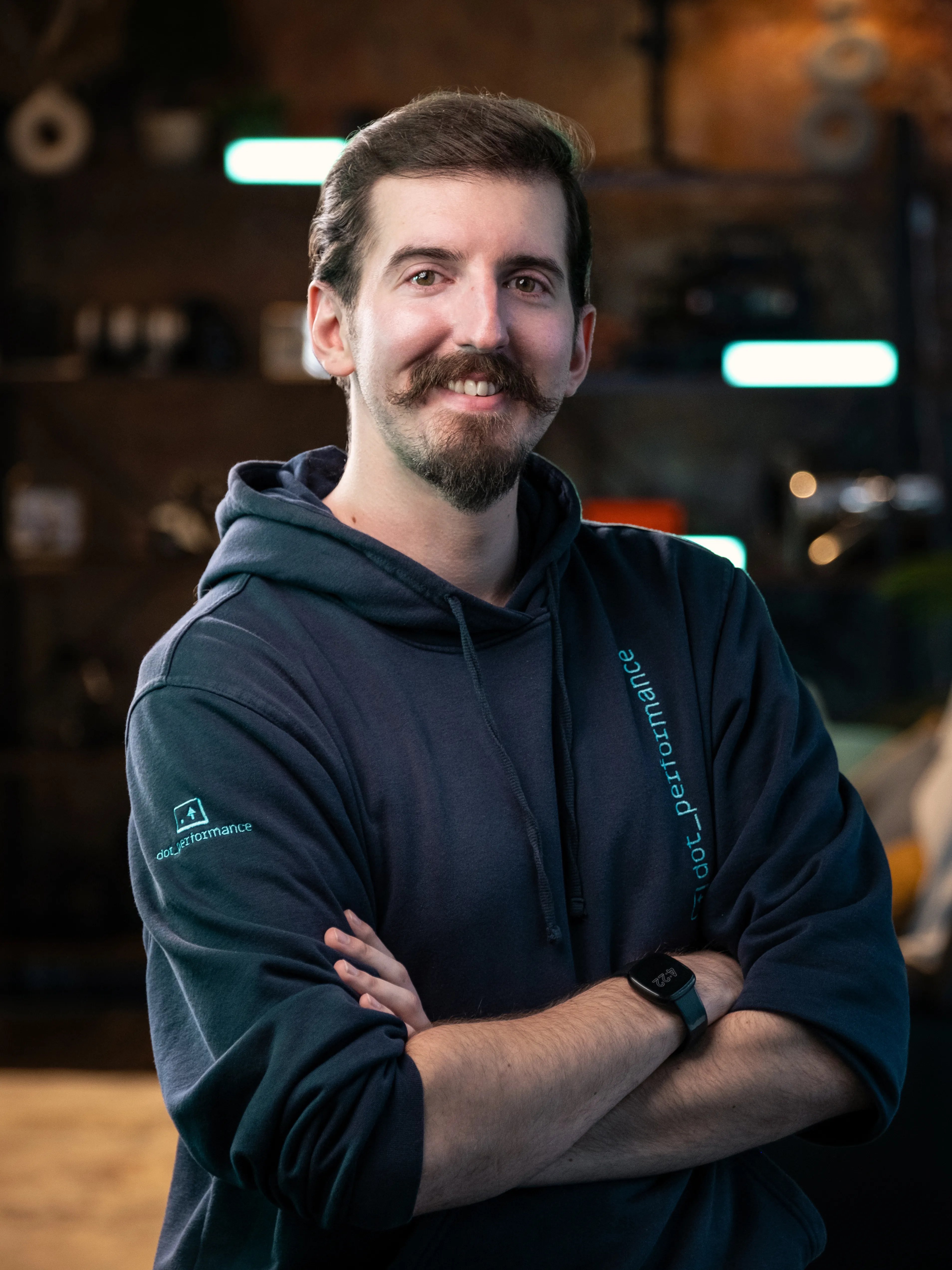 Will, cinematographer and editor at Dot Performance, smiling with crossed arms as he poses for headshot wearing branded hoody, against dark background with blue neon lights.