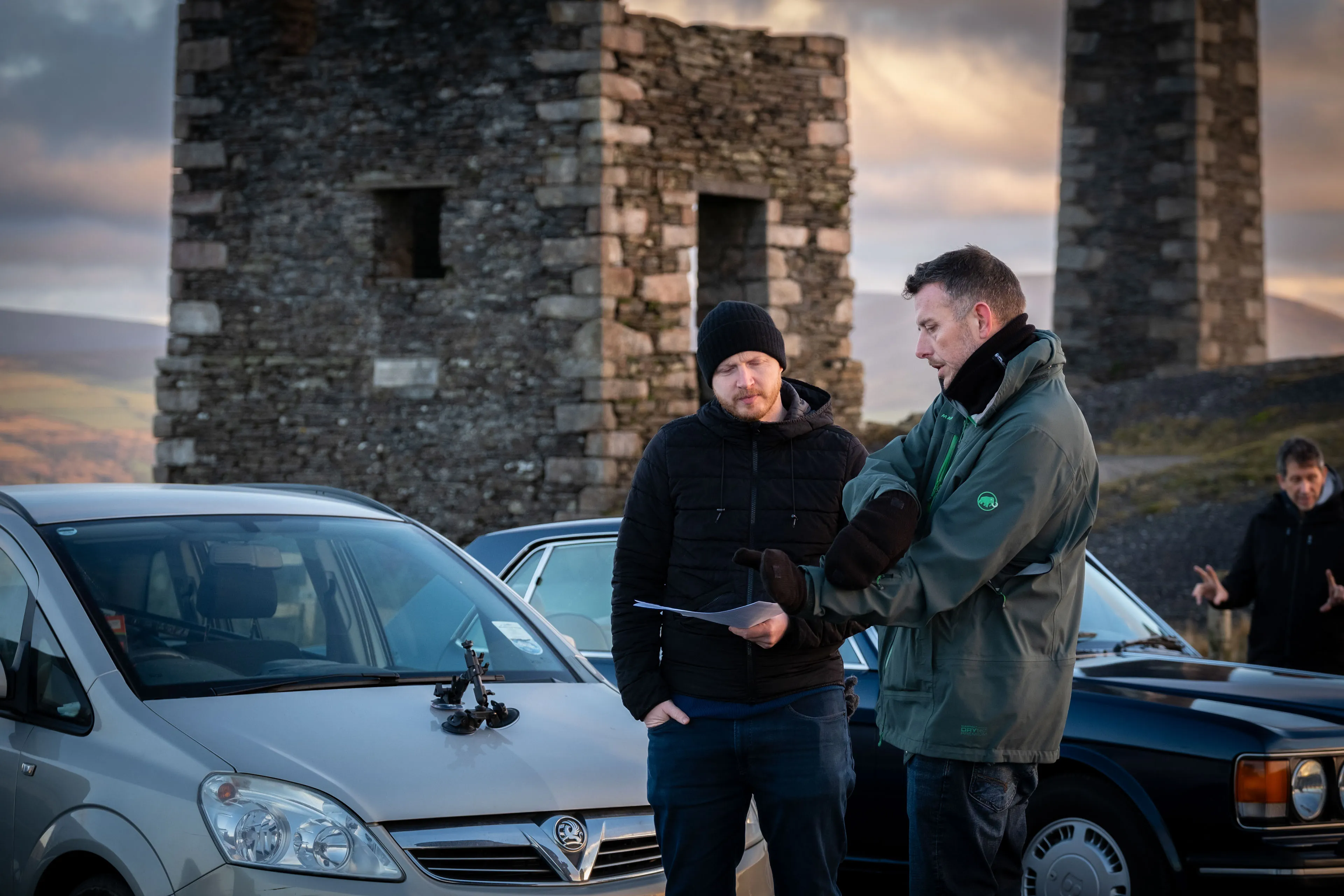 Two men stand by a silver car with a clipboard, wearing winter clothing. Stone ruins are in the background under a moody, cloudy sky.