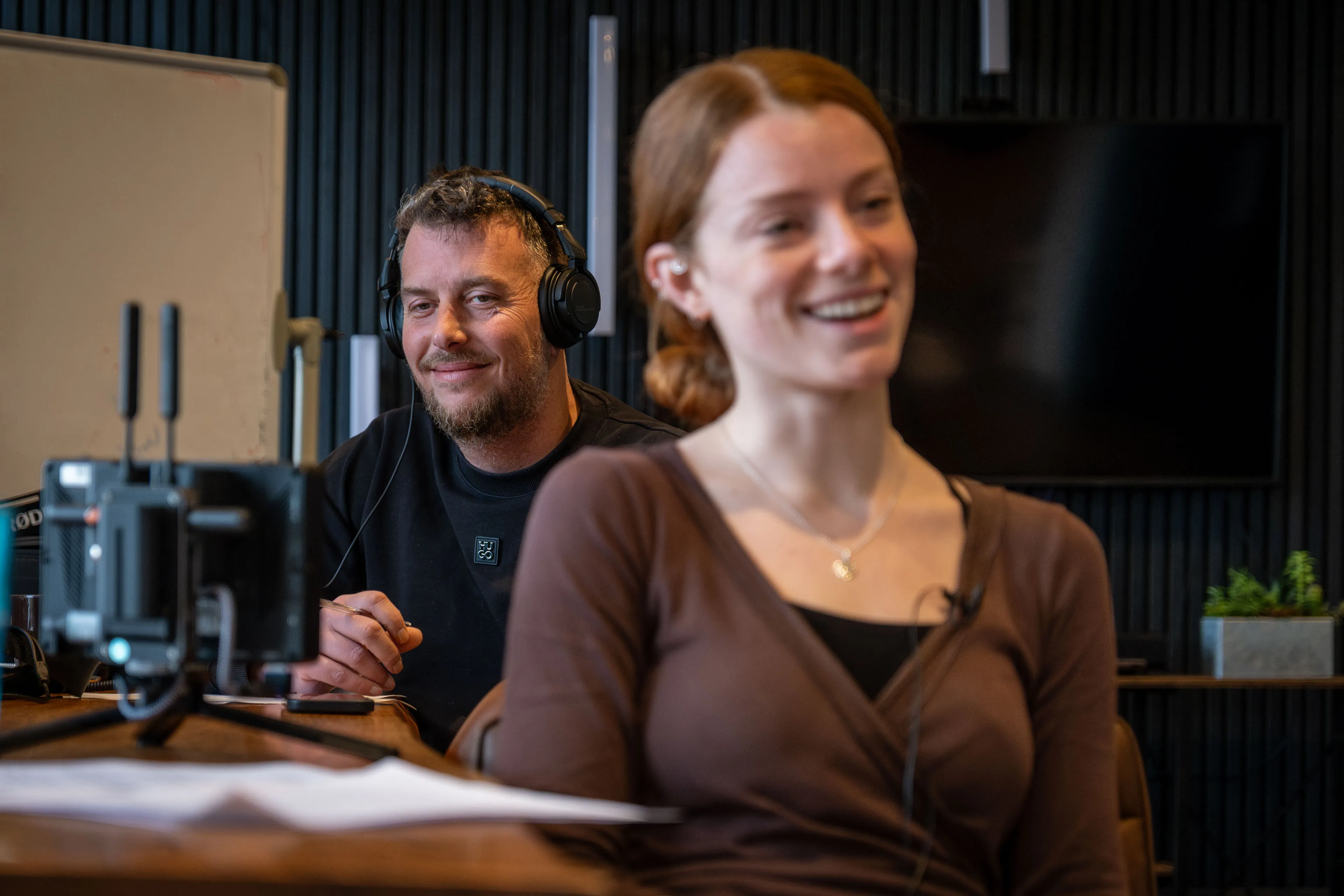 A smiling man wearing headphones and a woman in focus, sitting at a table with equipment in a modern office setting, creating a relaxed atmosphere.