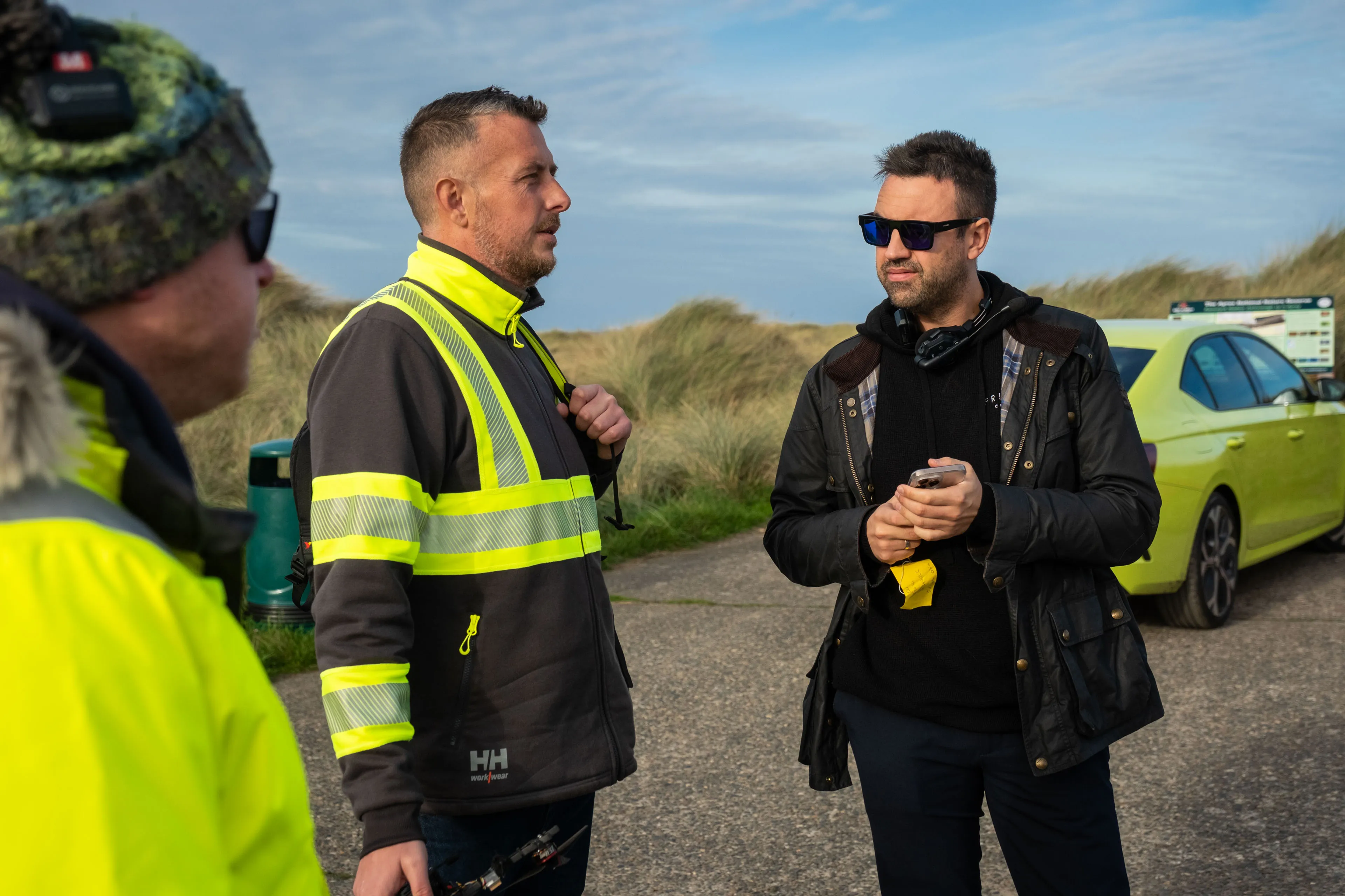 Three men stand on a dune-filled road beside a lime green Skoda car. The sky is clear and the mood appears focused and engaged.