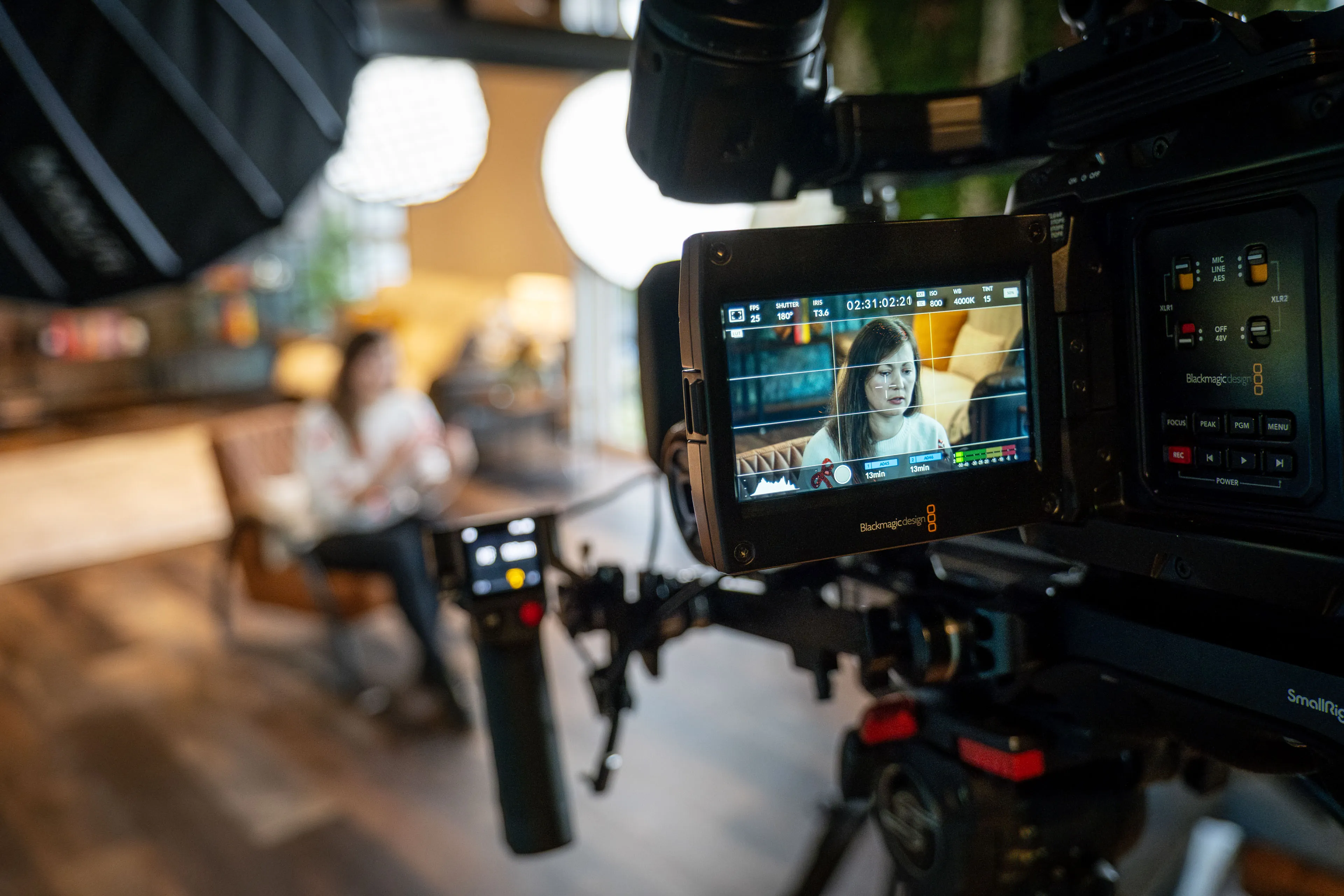 A video camera records a woman seated in a well-lit room, displayed on the camera's screen. The setting feels warm and professional.
