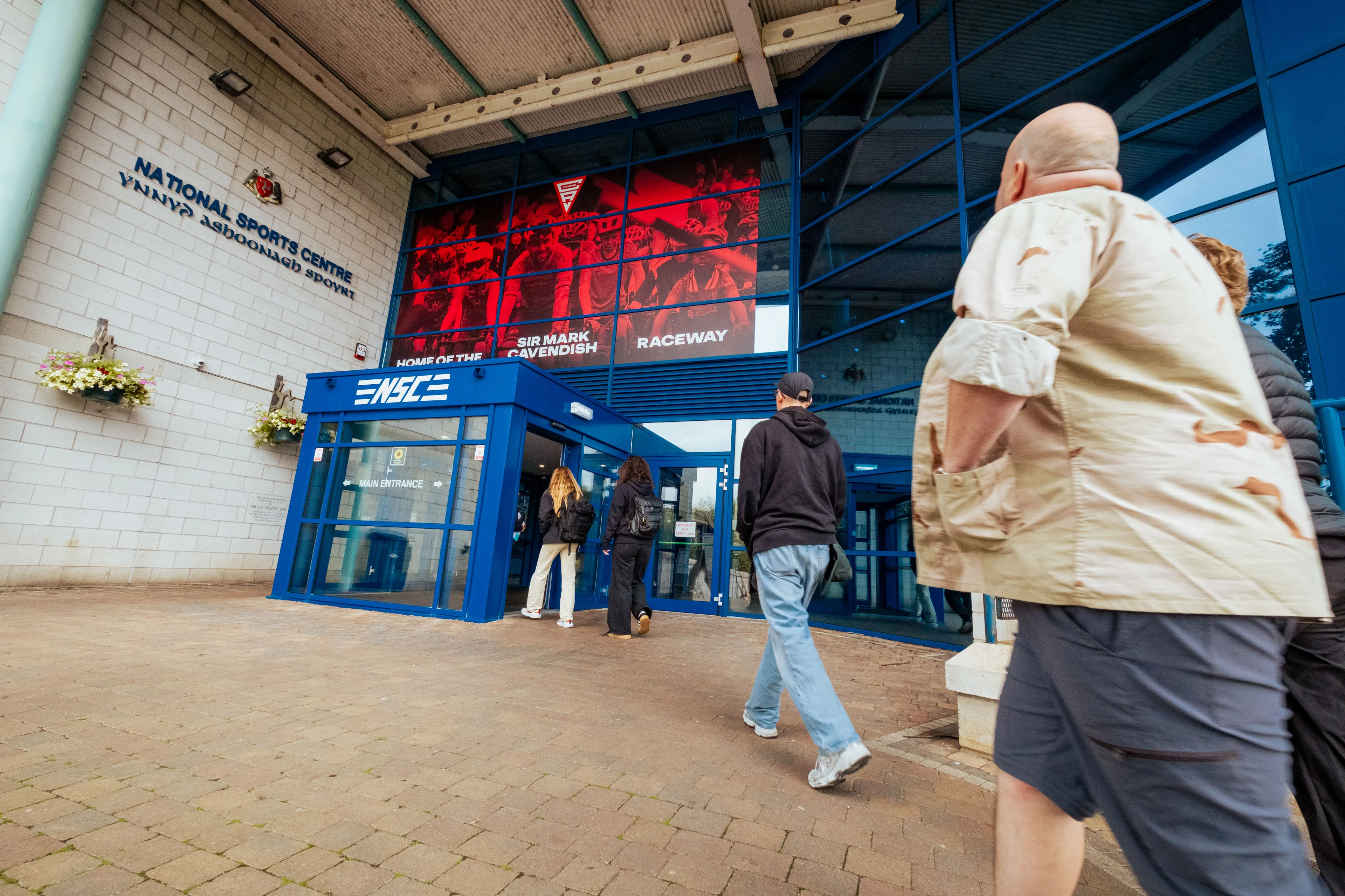 People entering the National Sports Centre through a blue entrance. A large red banner with race imagery is overhead. The scene is lively and welcoming.