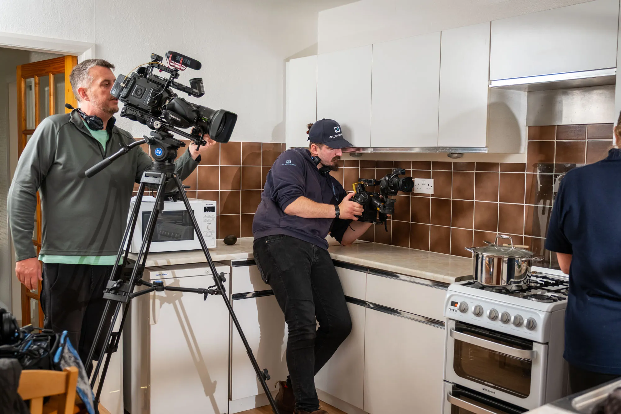 Two cameramen film in a kitchen with brown tiles and white cabinets. One operates a tripod camera; the other holds a camera near a stove, focusing intently.