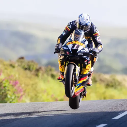 Motorcyclist in colorful gear performs a wheelie on a racing bike labeled "Milenco" on a sunlit road, conveying excitement and skill.