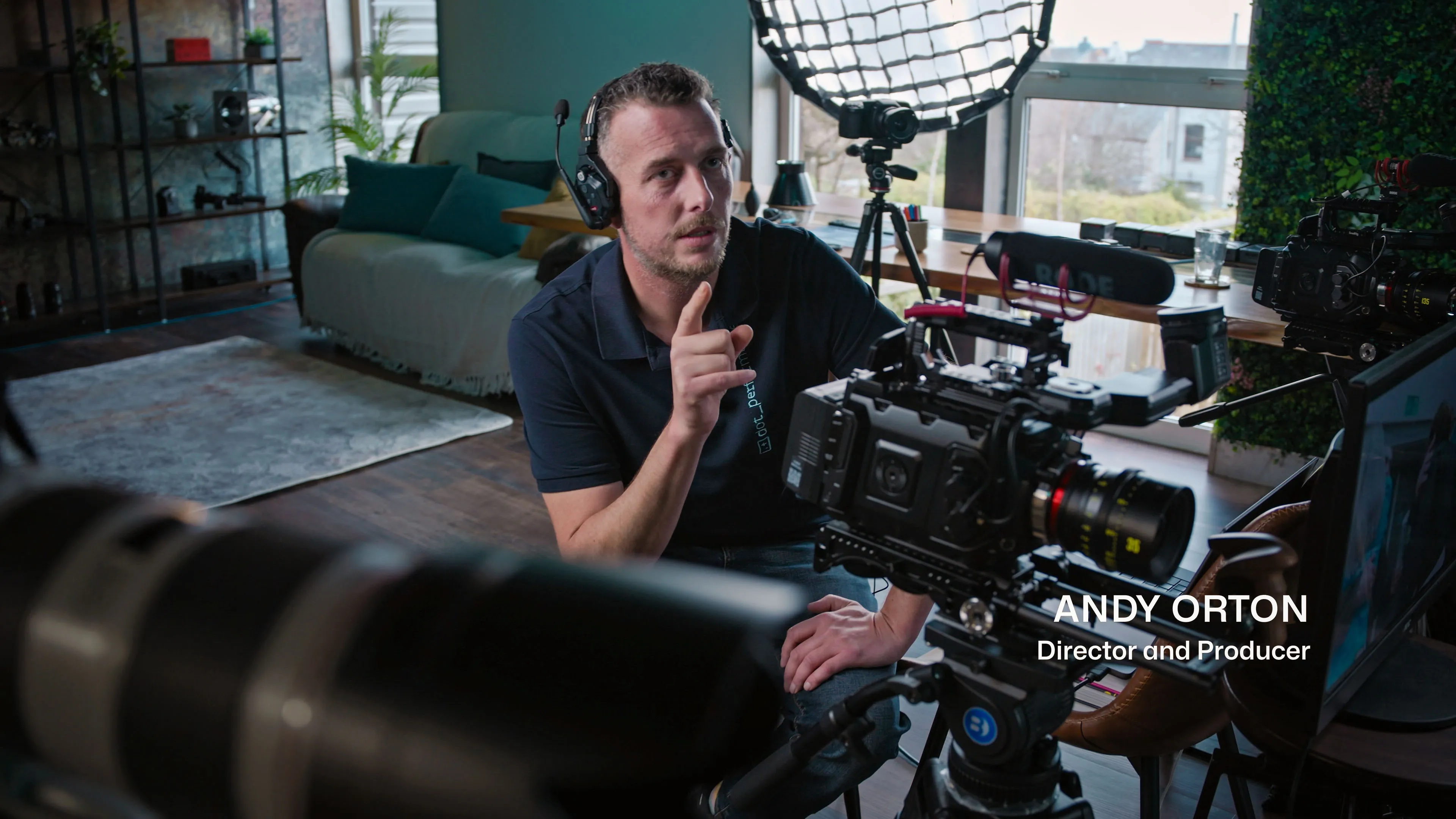 Man wearing headphones, seated in a studio setting surrounded by film equipment. He appears focused, gesturing with one hand. Calm and professional atmosphere.