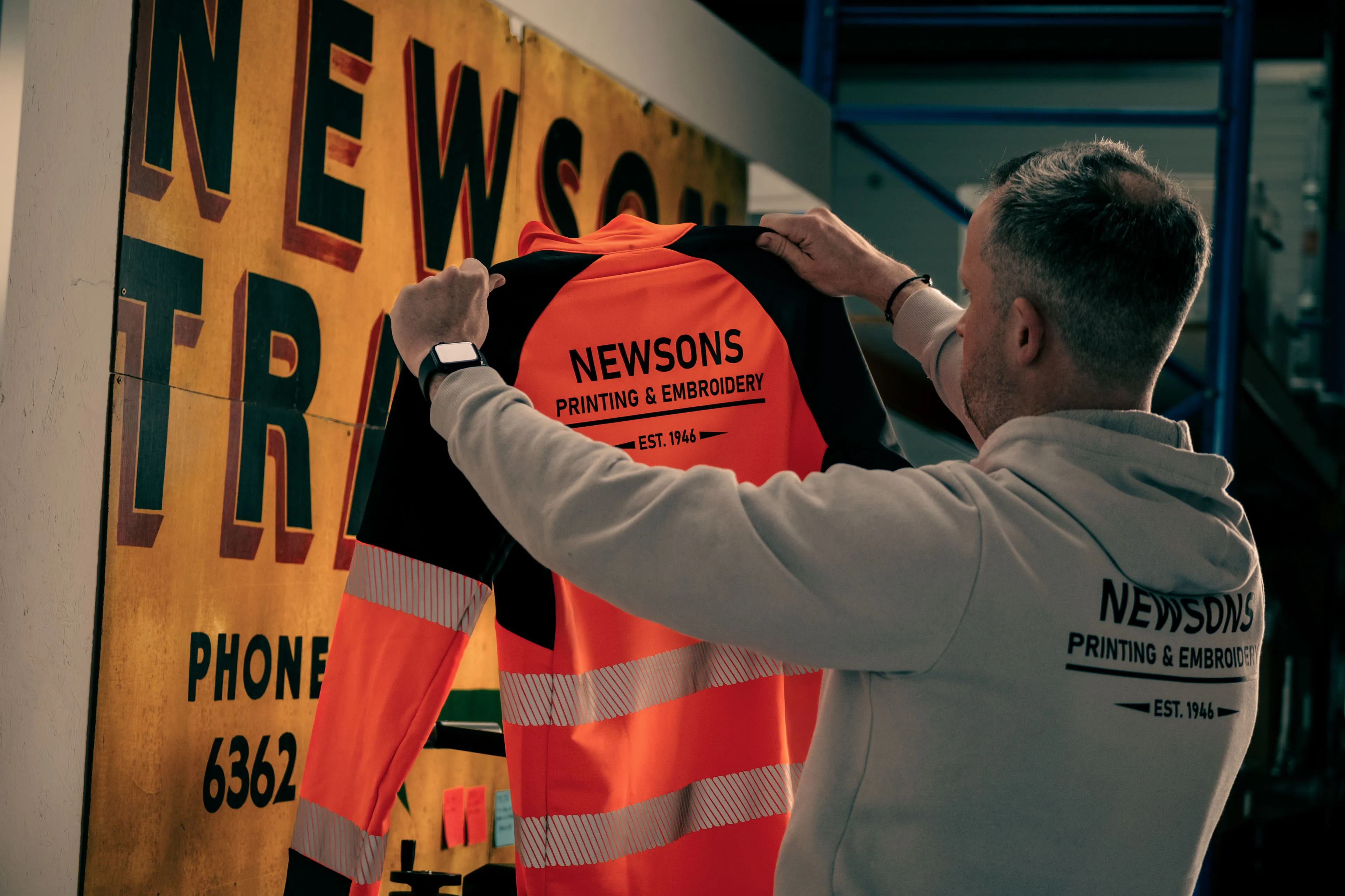 Man holding a high-visibility orange work shirt with "Newsons Printing & Embroidery" text. Background features vintage signage, creating a nostalgic tone.