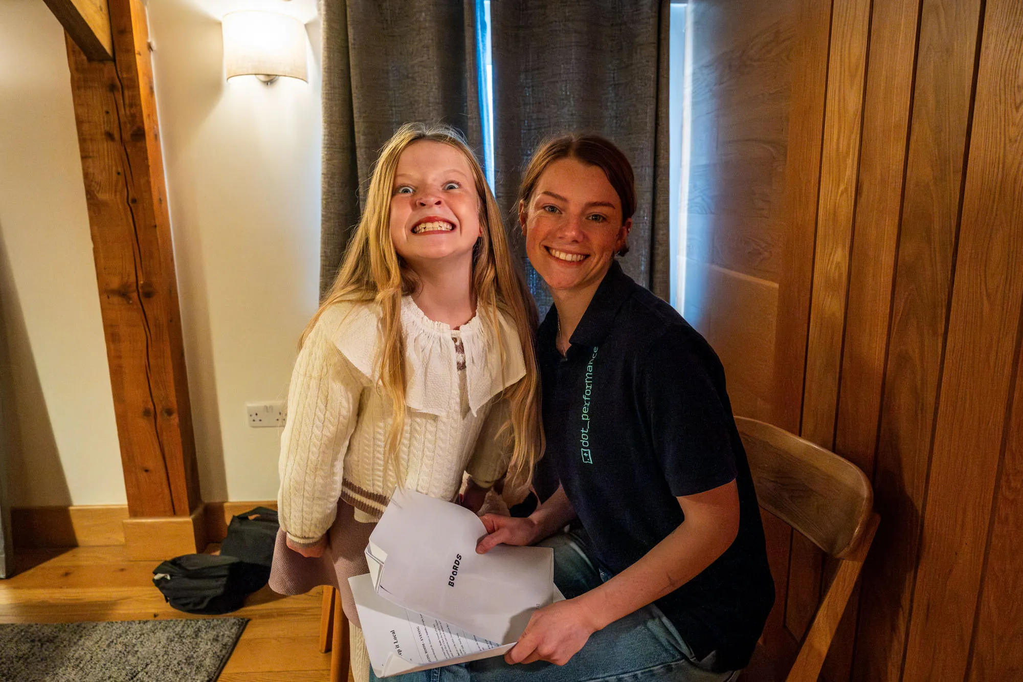 A young girl and a woman smiling brightly indoors. The girl wears a white sweater while the woman holds papers, sitting on a wooden chair, exuding warmth and happiness.