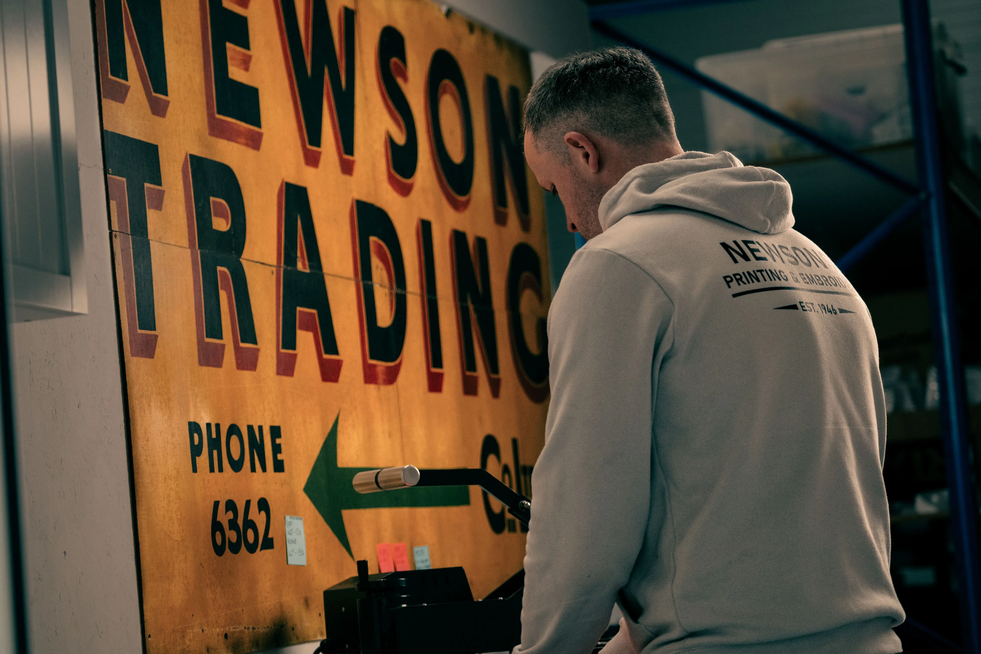 A person in a hoodie is standing near a vintage sign reading "Newson Trading" in a dimly lit room. The atmosphere feels industrial and nostalgic.
