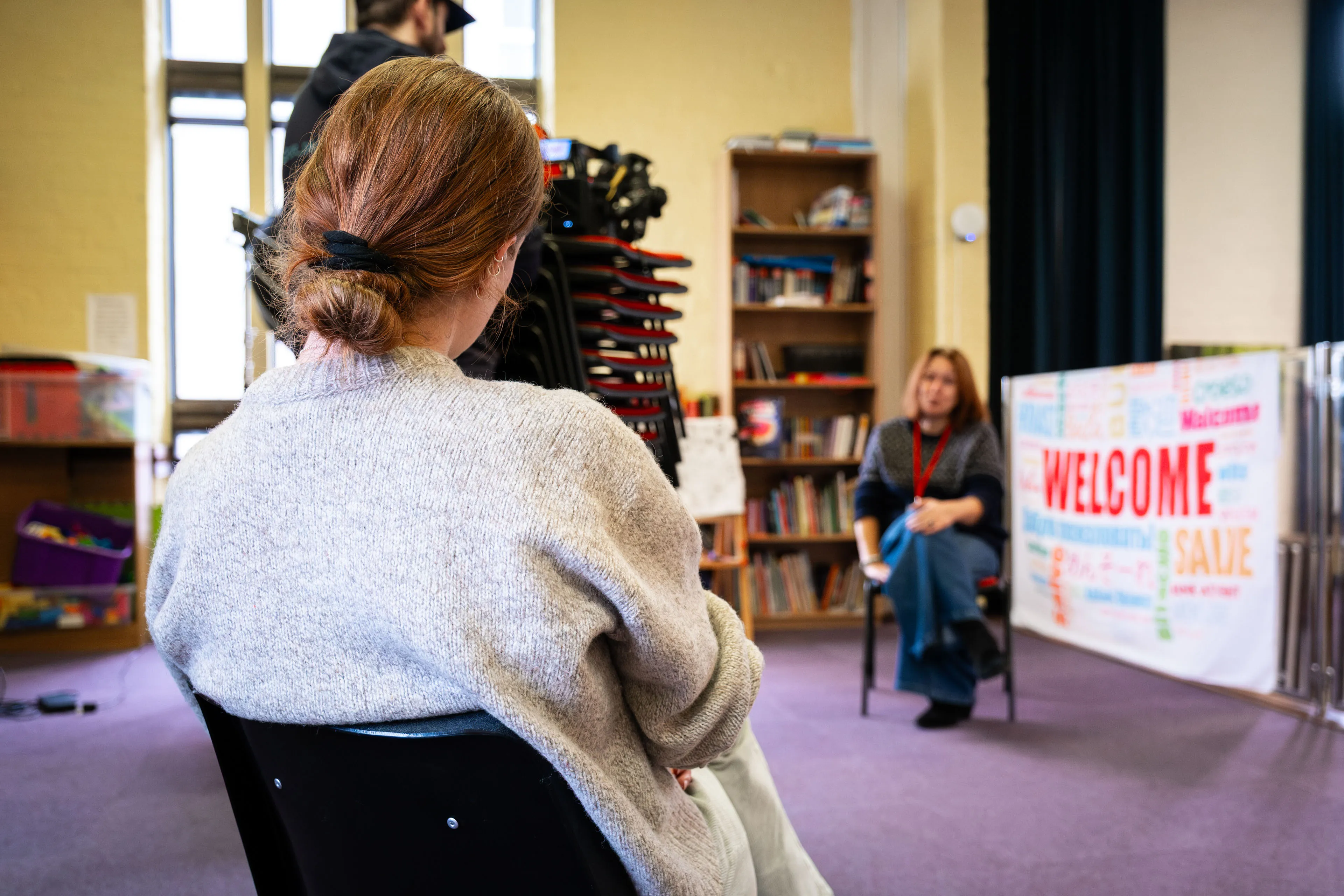 A woman with a ponytail sits in a classroom with bookshelves and a "WELCOME" poster. A person with a camera films the scene, creating an atmosphere of anticipation.