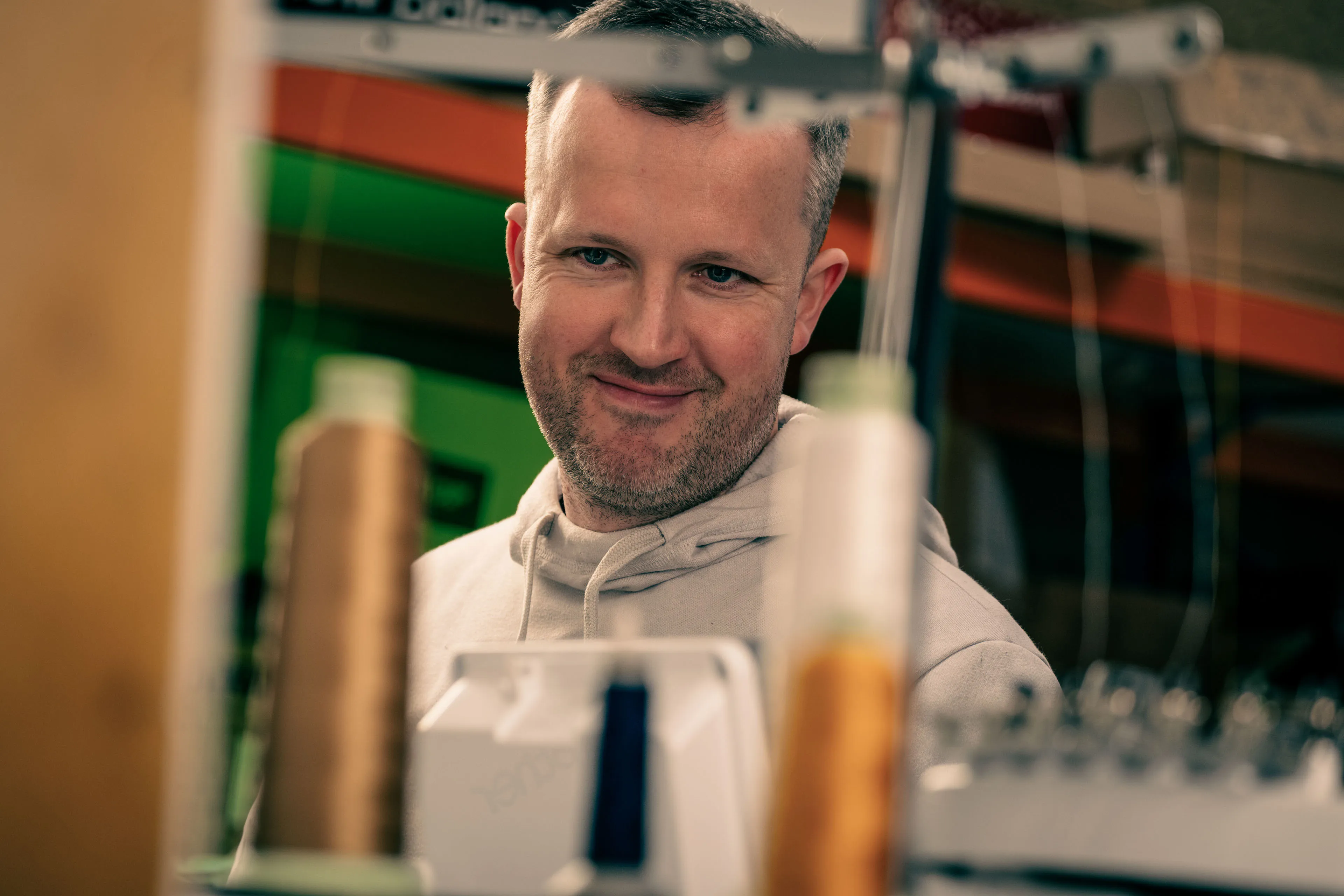 A man in a beige hoodie is smiling while looking at sewing equipment. Spools of thread are visible in the foreground, creating a warm, focused atmosphere.