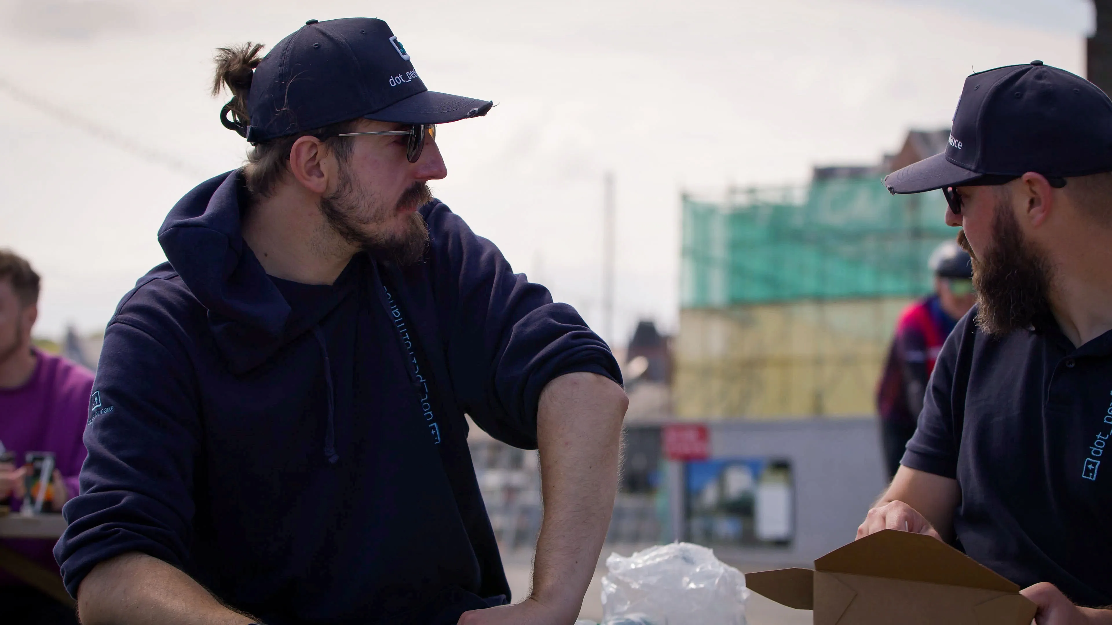 Two men wearing caps and dark hoodies sit outdoors, engaged in conversation. One has a beard; the setting appears casual with buildings in the background.