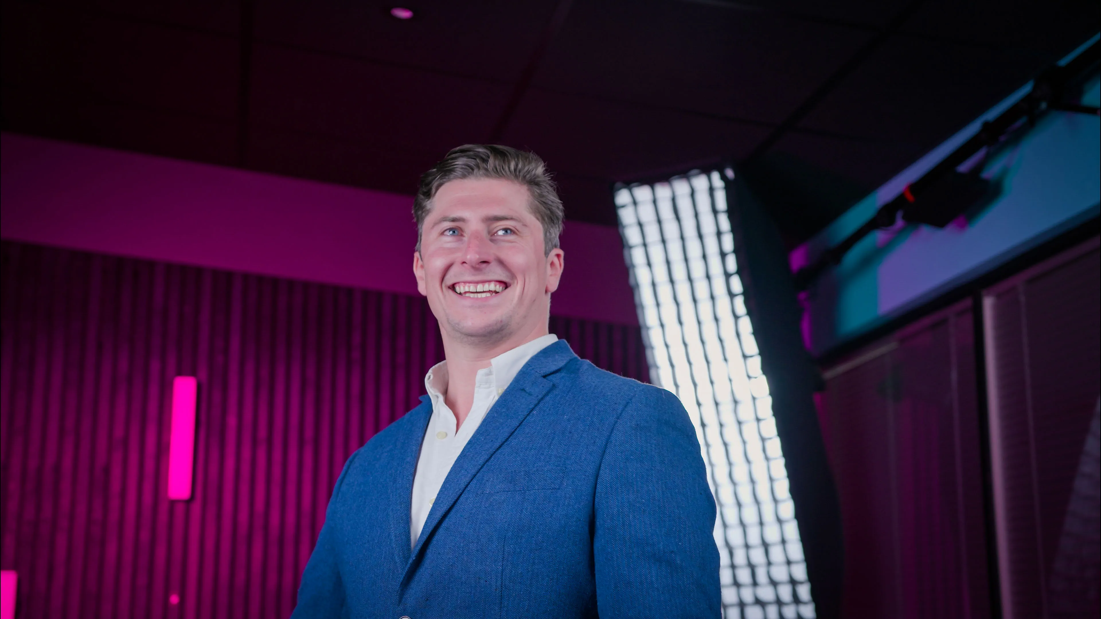 Smiling man in a blue blazer stands in a studio with vibrant pink and purple lighting. The ambiance is modern and energetic, conveying a joyful mood.