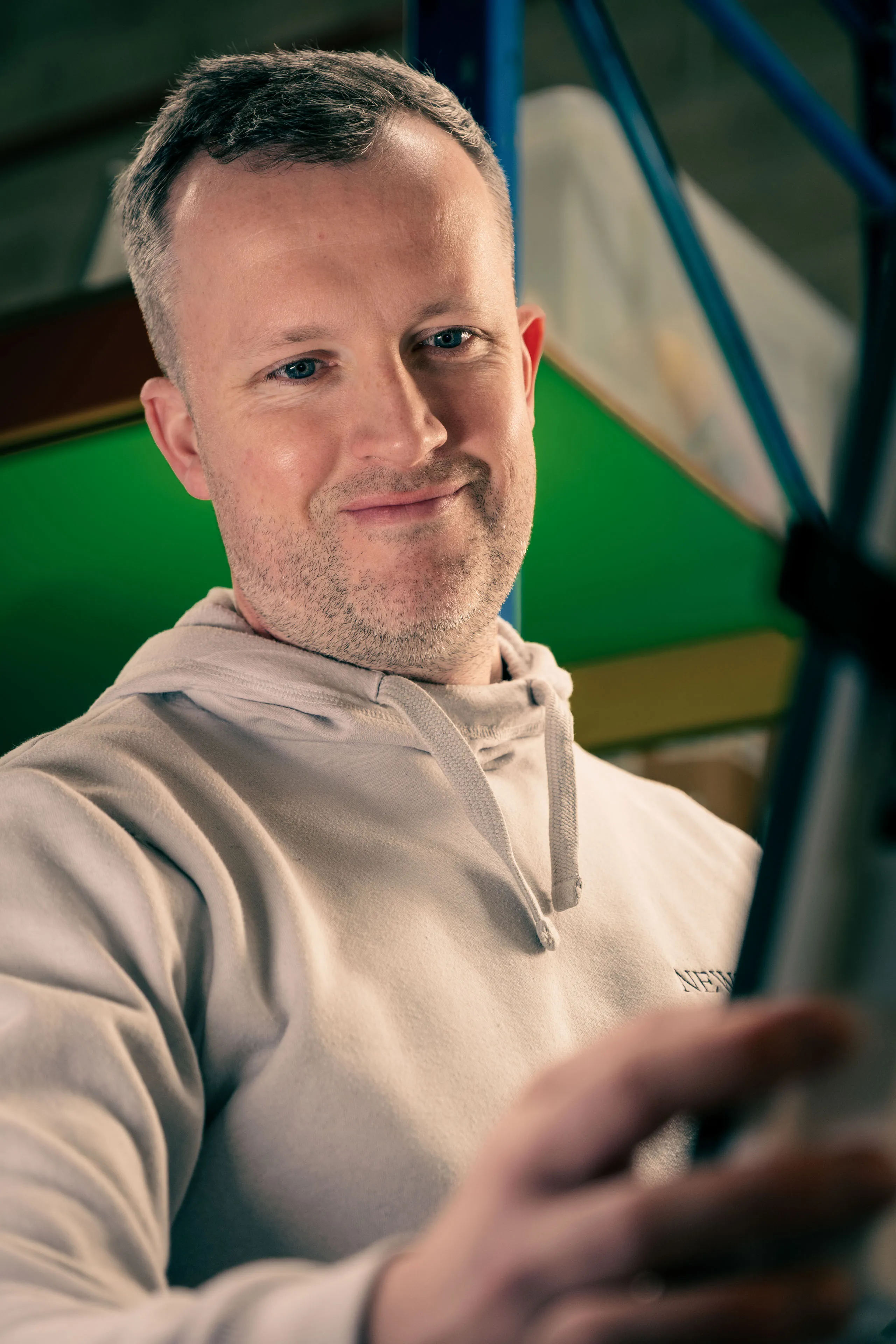 A man in a light hoodie smiles gently, working on a machine's computer display. The background is vibrant with green and yellow hues, creating a warm, approachable mood.