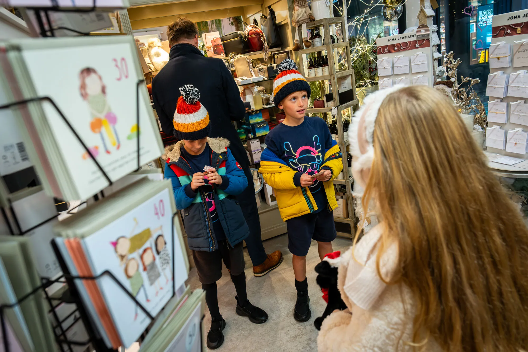 Two children in colourful winter jackets and hats browse a shop filled with cards and gifts. A girl with long hair stands nearby, creating a cheerful and festive atmosphere.
