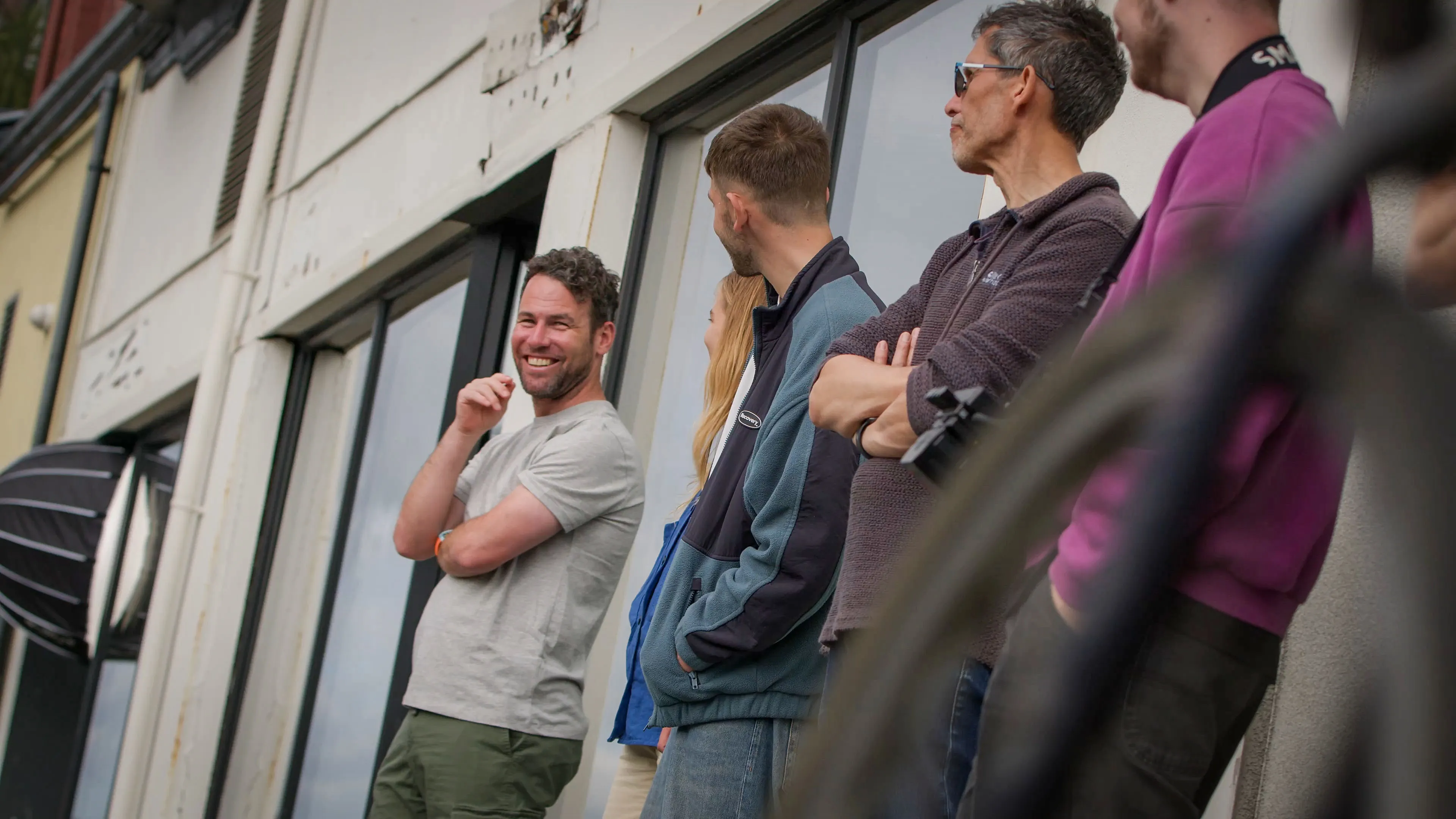 A group of people stand casually outside a building, smiling and chatting. One man in a grey shirt is laughing, creating a friendly and relaxed atmosphere.