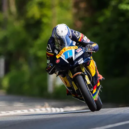 Motorcyclist in full gear rides a racing bike with "Milenco" branding, setting a dynamic tone. Background shows a blurred, green, tree-lined street.