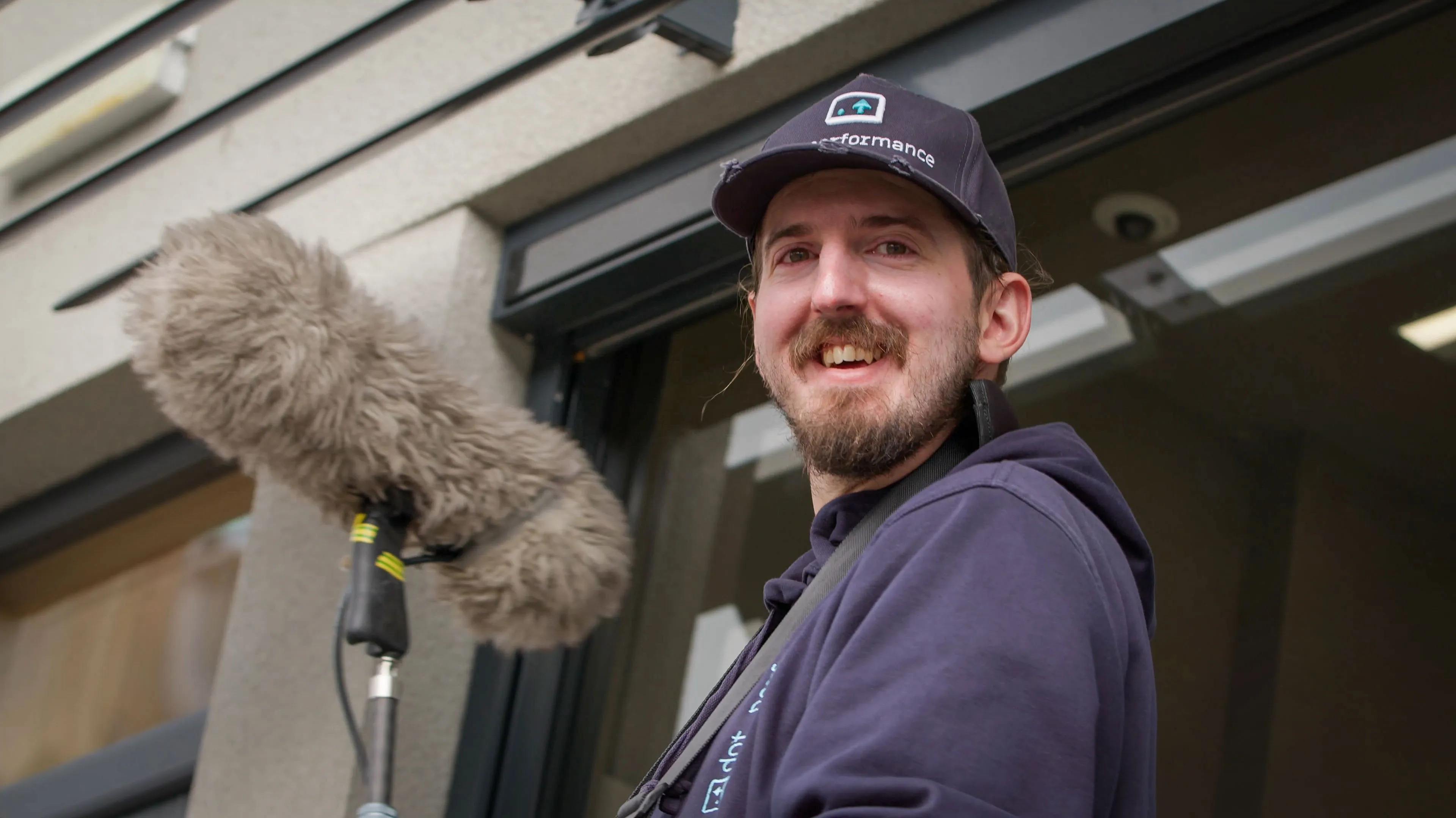 A man wearing a cap and hoodie smiles warmly while holding a boom microphone outside a building. The scene conveys a friendly, professional vibe.