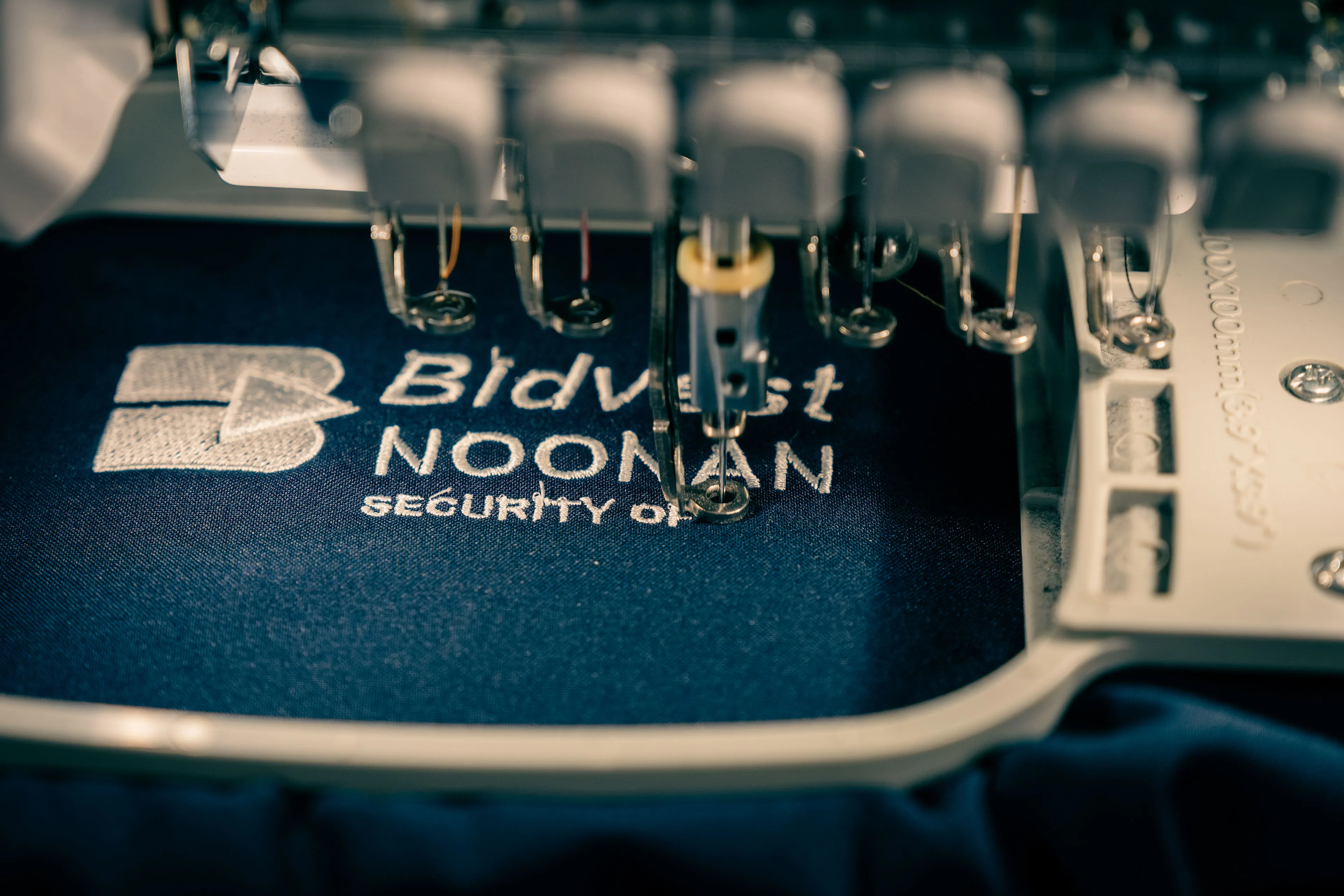 Close-up of an embroidery machine stitching "Bidvest Noonan Security" logo in white thread on dark fabric. Mechanical precision and textile detail.
