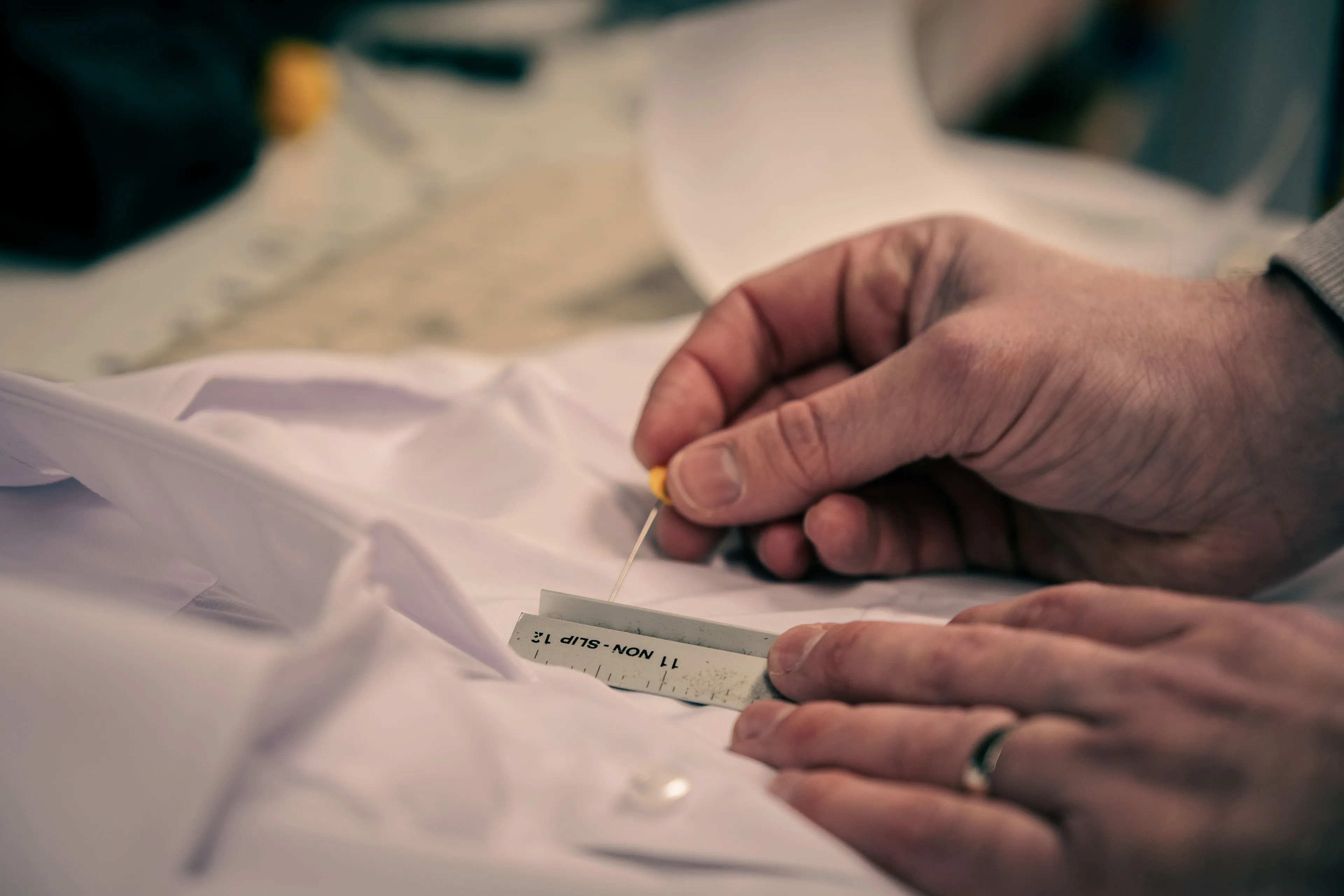 Hands carefully pinning a tag to a crisp, white shirt on a table. The scene conveys precision and craftsmanship in clothing preparation.