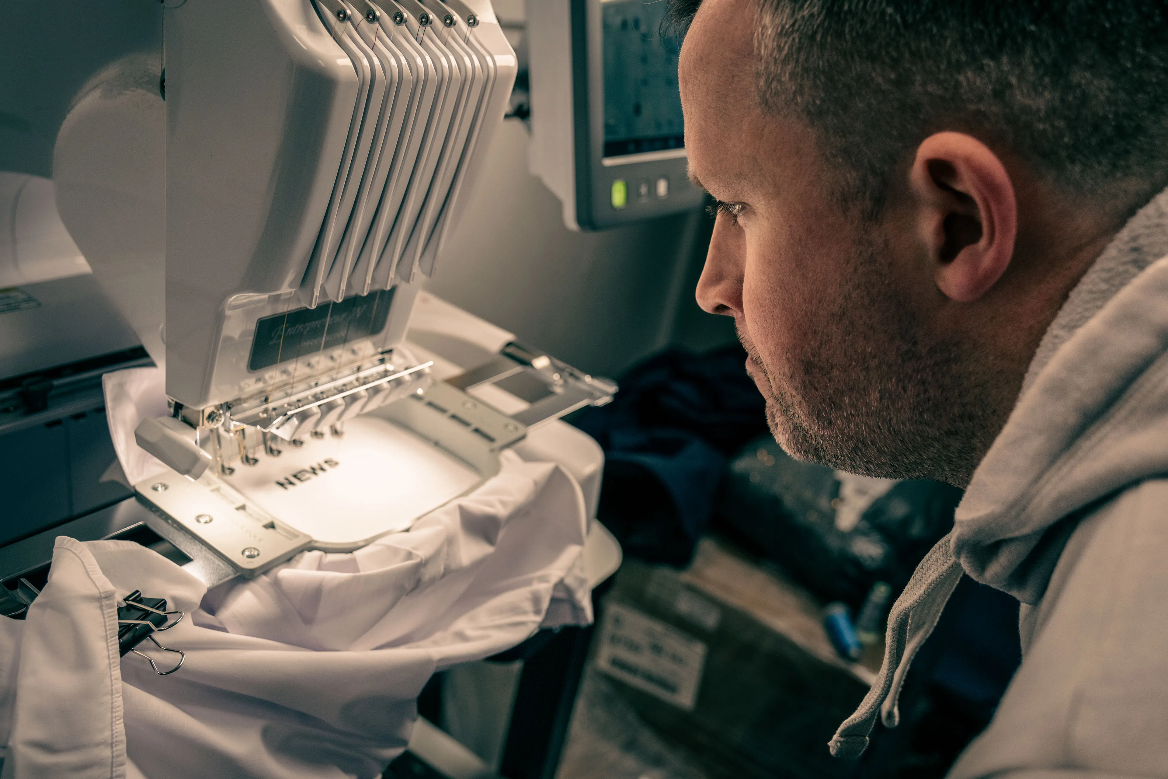 A man intently oversees an embroidery machine stitching text on white fabric. The setting is warmly lit, highlighting focused craftsmanship.