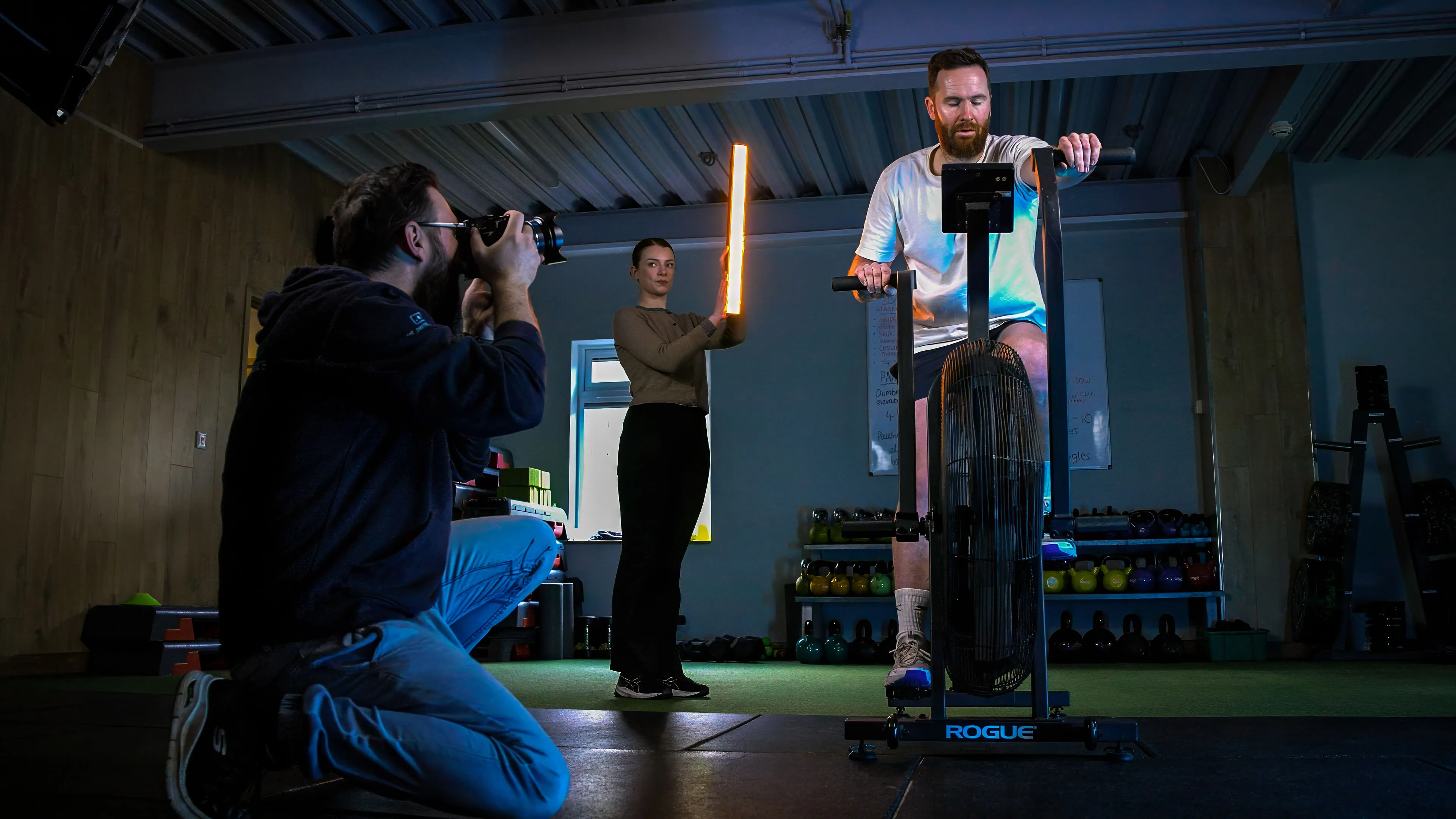 A man intensely rides an air bike in a gym while a photographer captures the moment. A woman holds an LED light, emphasising a focused atmosphere.